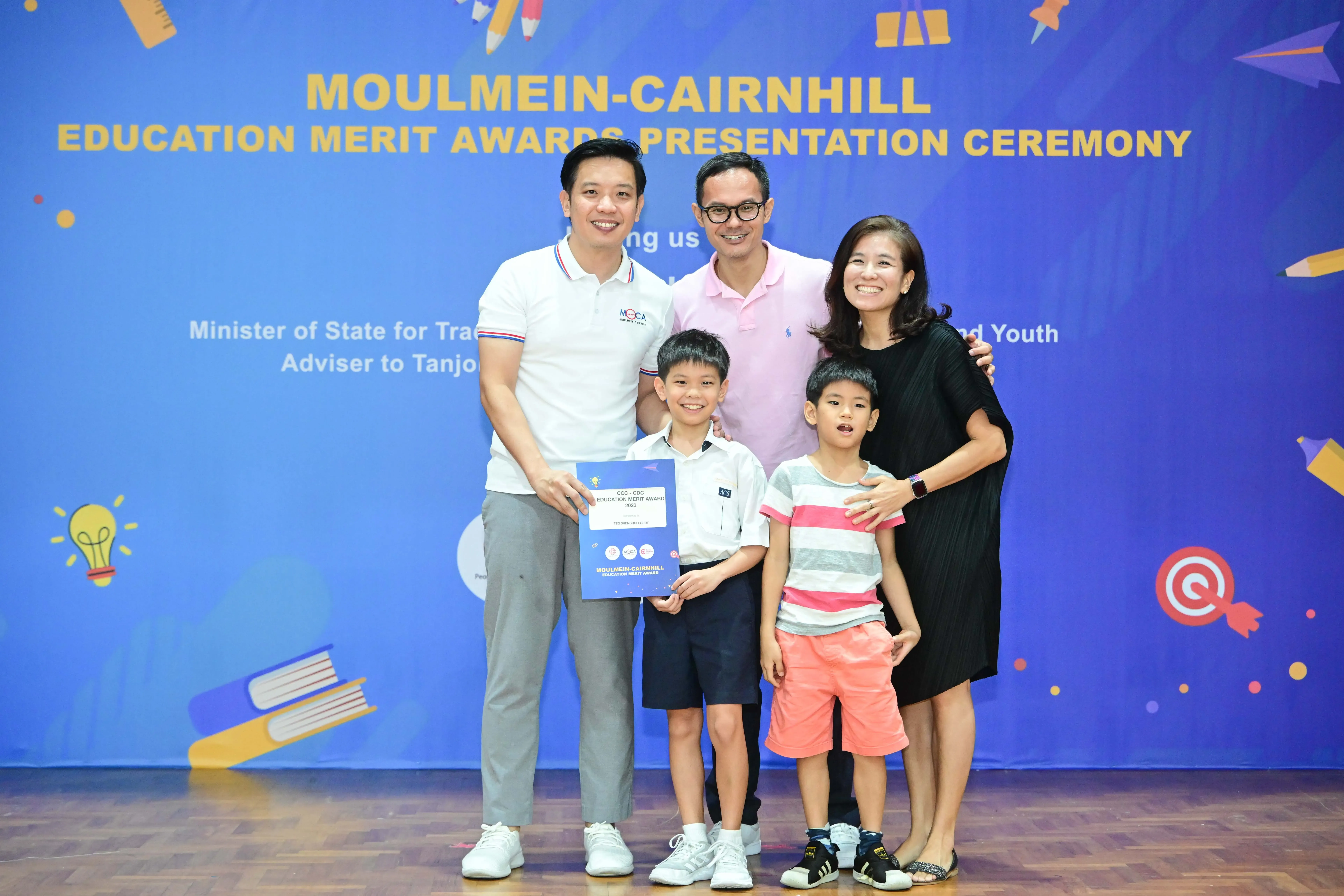 Five people pose before a Moulmein-Cairnhill Education Merit Awards banner, with a boy holding an award certificate.