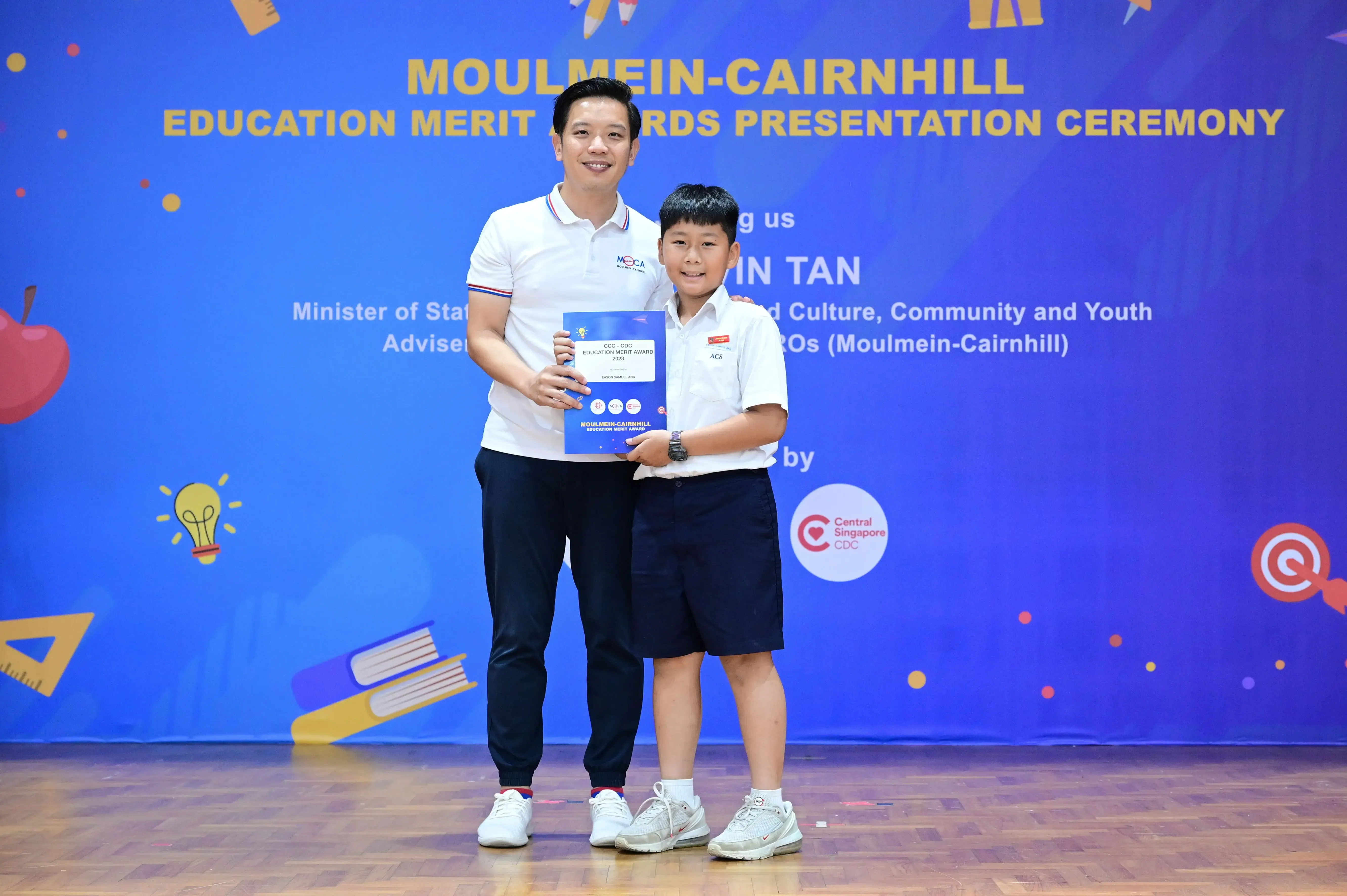 An adult and a young boy hold an award certificate on stage at the Moulmein-Cairnhill Education Merit Awards Ceremony.