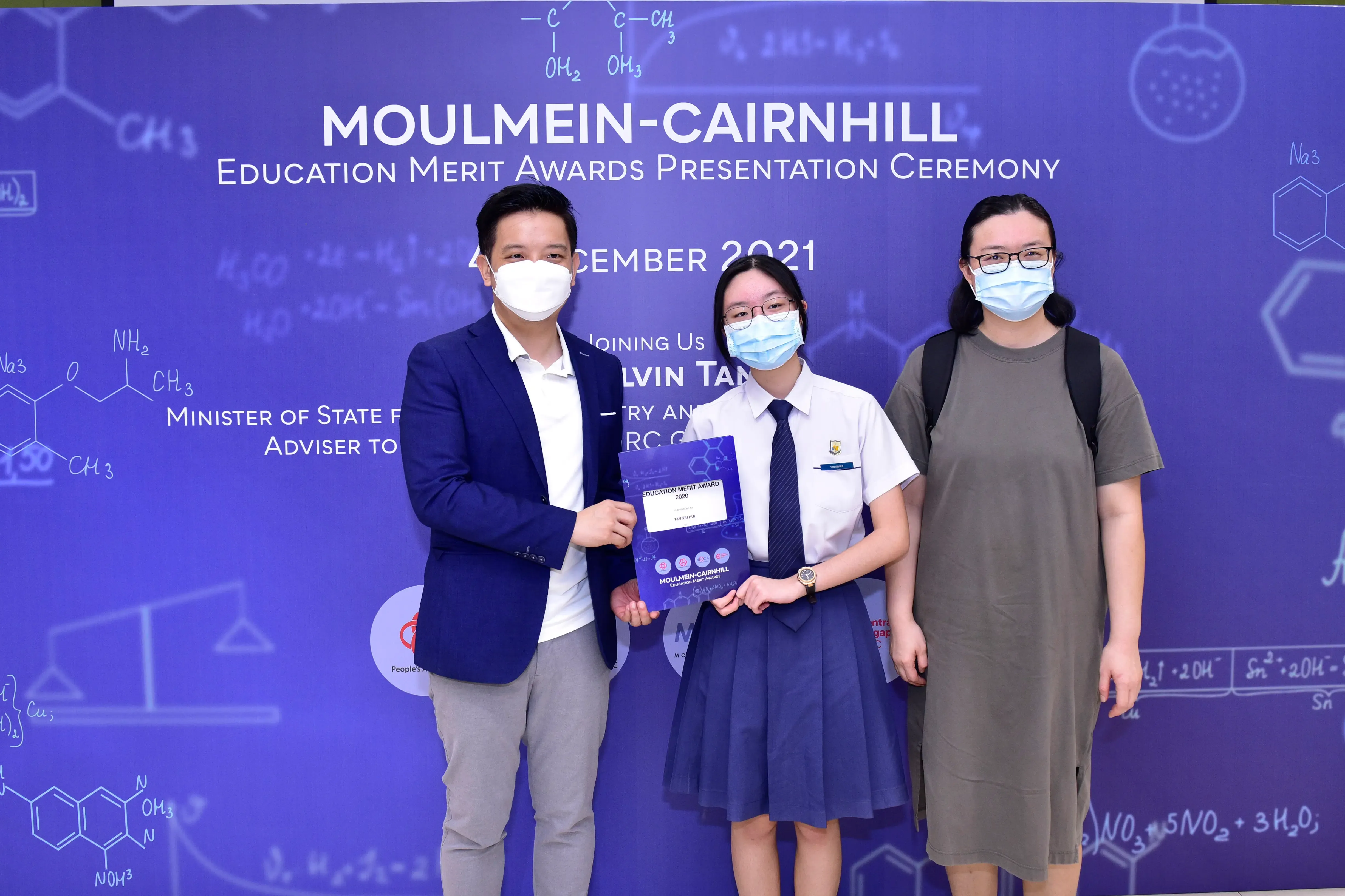Three masked people pose at the Moulmein-Cairnhill Education Merit Awards Ceremony 2021, with a student holding a certificate.