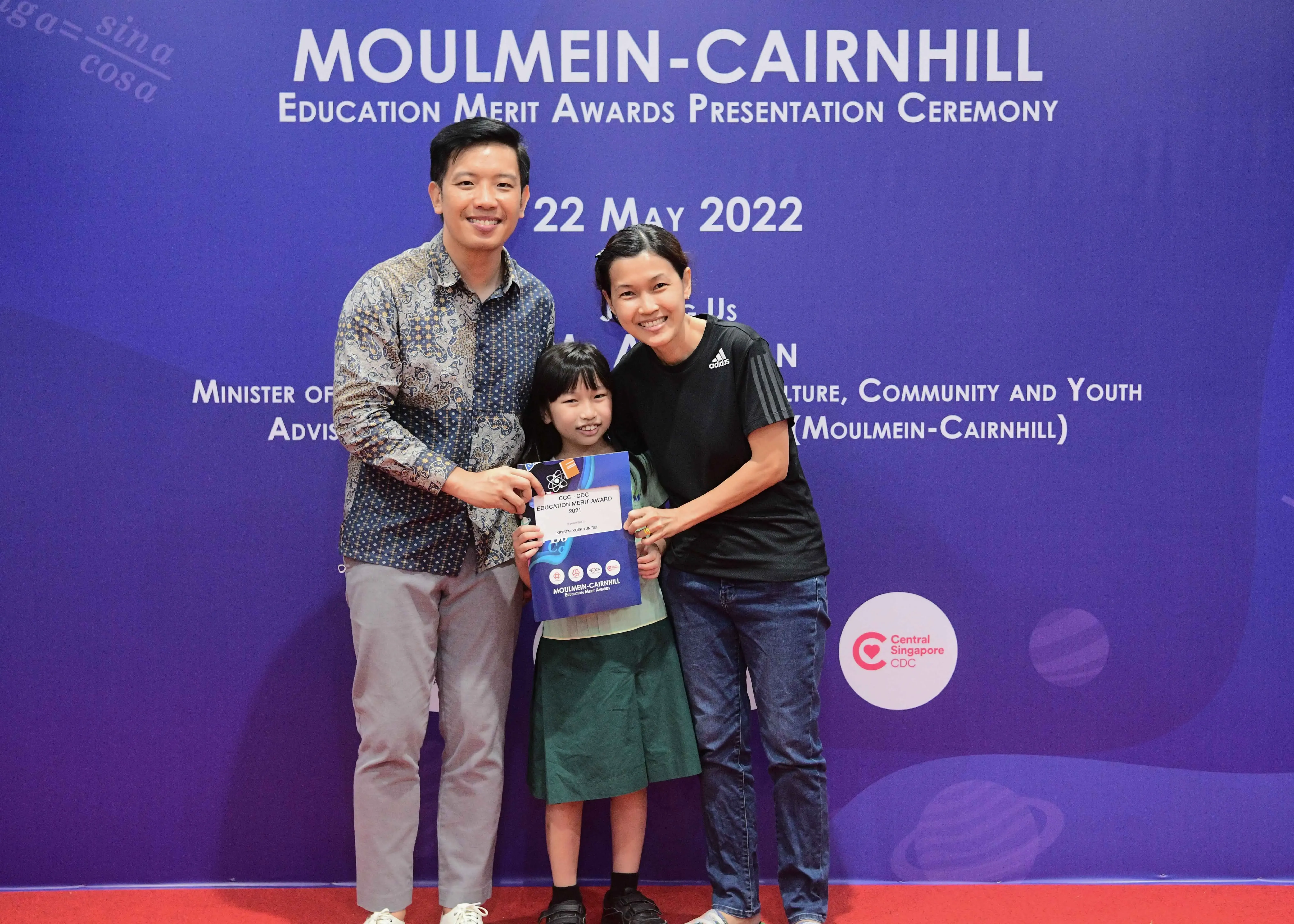 Two adults and a young girl pose with an award certificate at the Moulmein-Cairnhill Education Merit Awards ceremony on 22 May 2022.