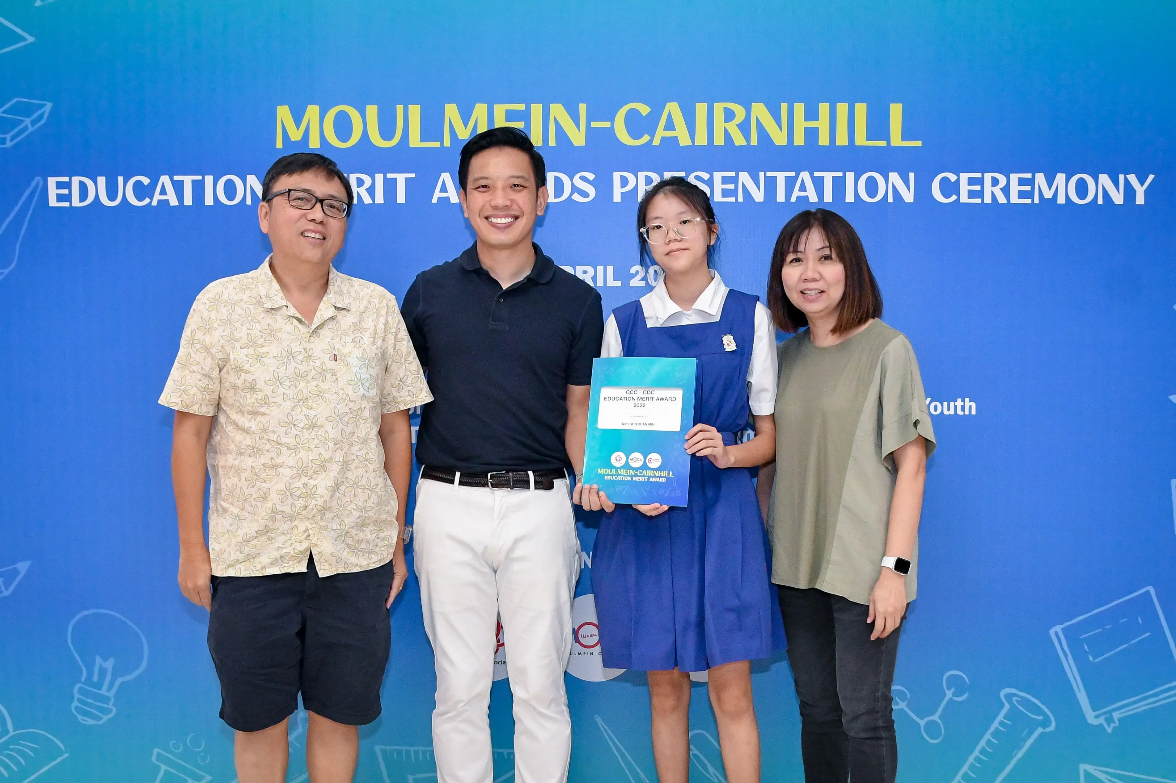 Four people pose before a Moulmein-Cairnhill Education Merit Awards banner, a girl in school uniform holding a certificate.