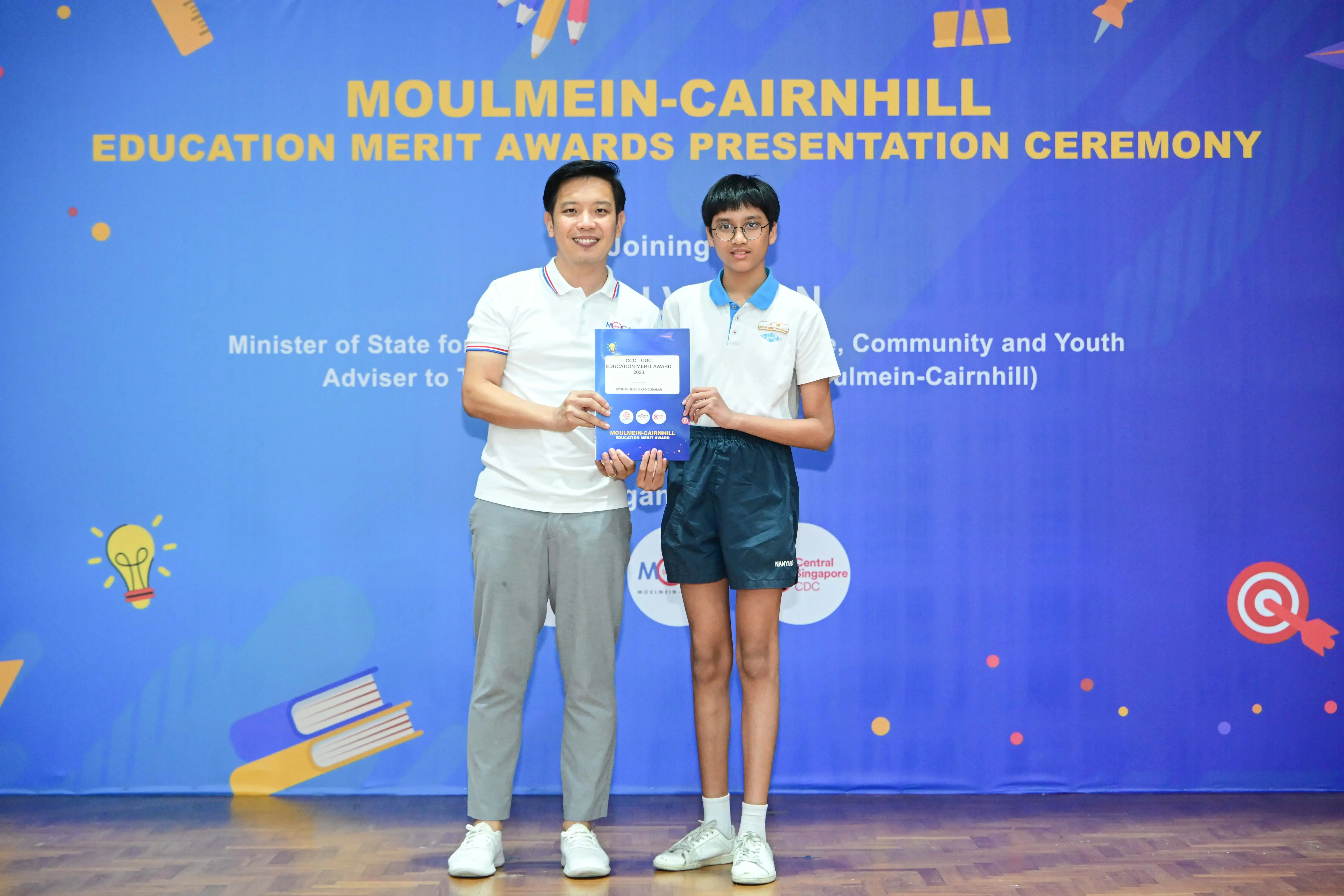 Two people hold a certificate in front of a Moulmein-Cairnhill Education Merit Awards Presentation Ceremony banner.