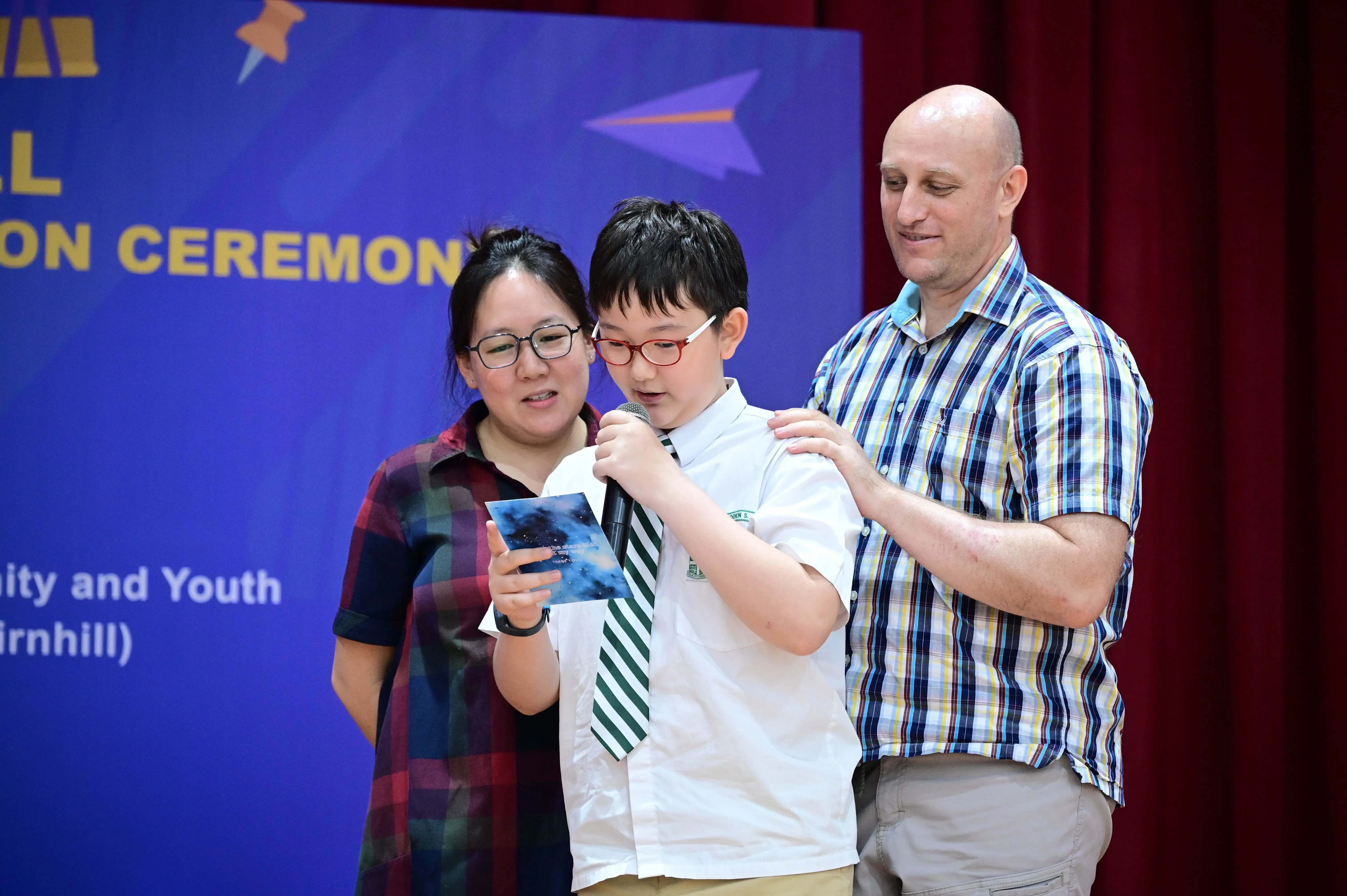 A boy in school uniform speaks into a microphone on stage, flanked by a woman and a bald man at a ceremony.