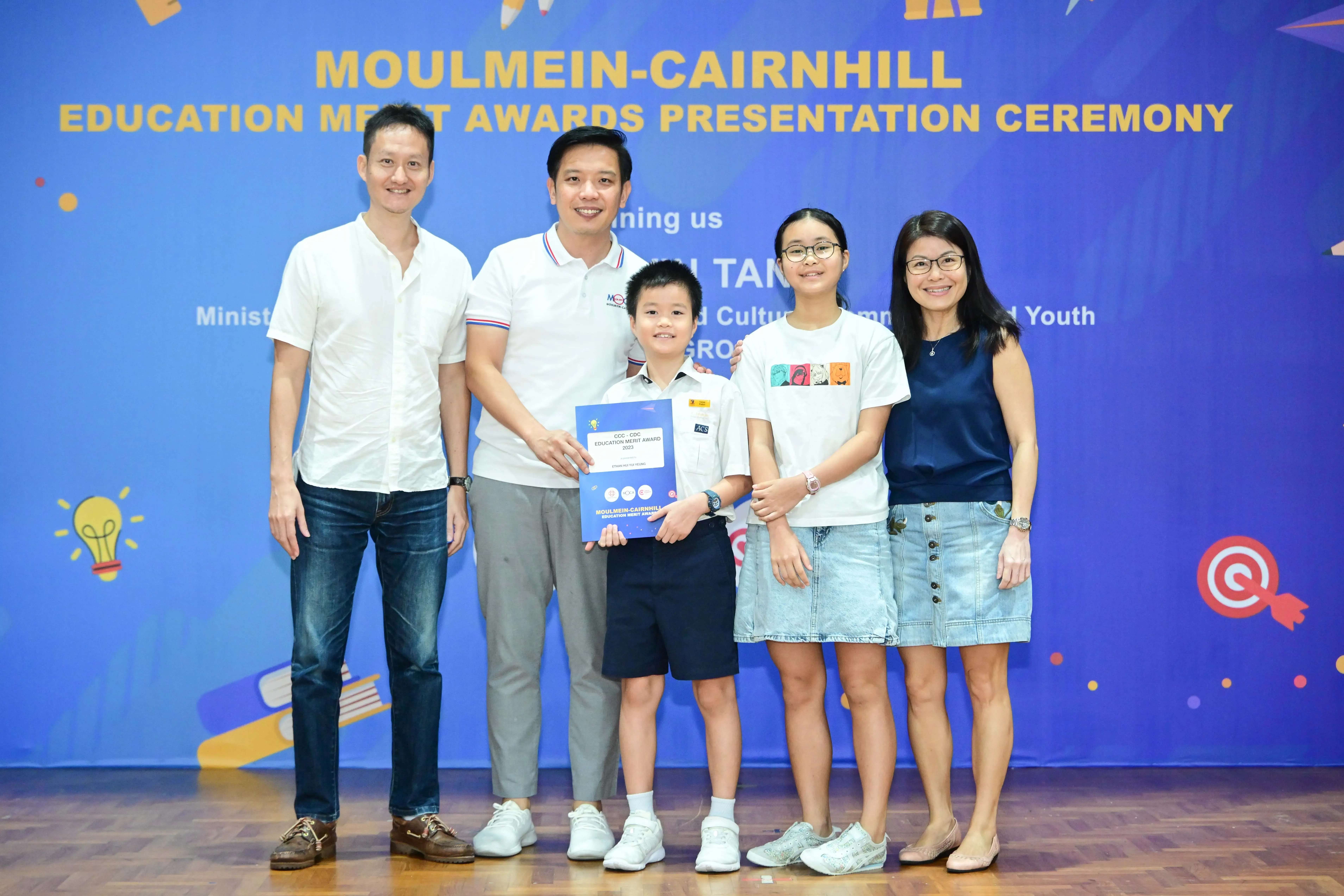 Five people pose at the Moulmein-Cairnhill Education Merit Awards Presentation Ceremony, with a boy holding a certificate.