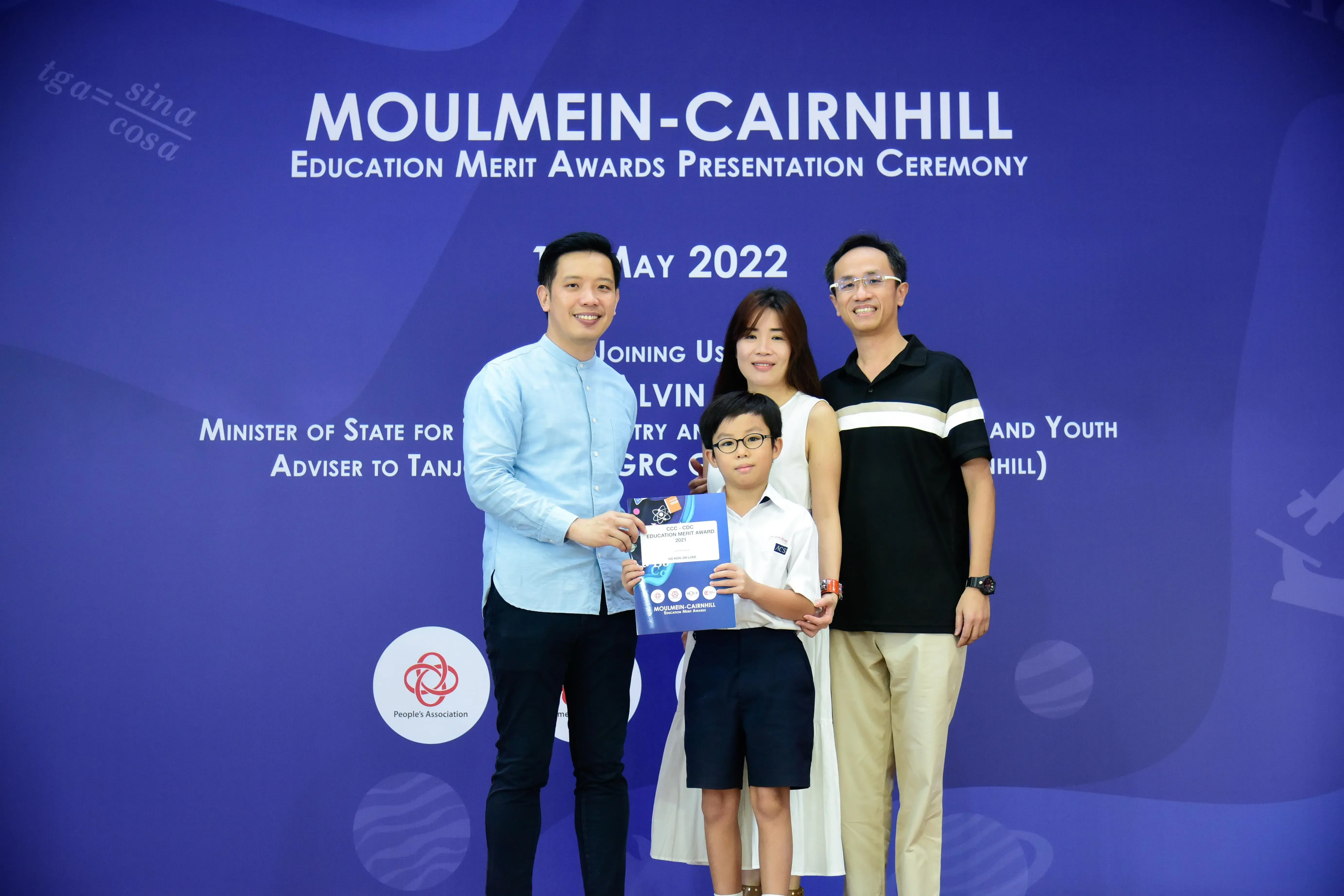 A man presents a certificate to a young boy flanked by two adults at the Moulmein-Cairnhill Education Merit Awards 2022.