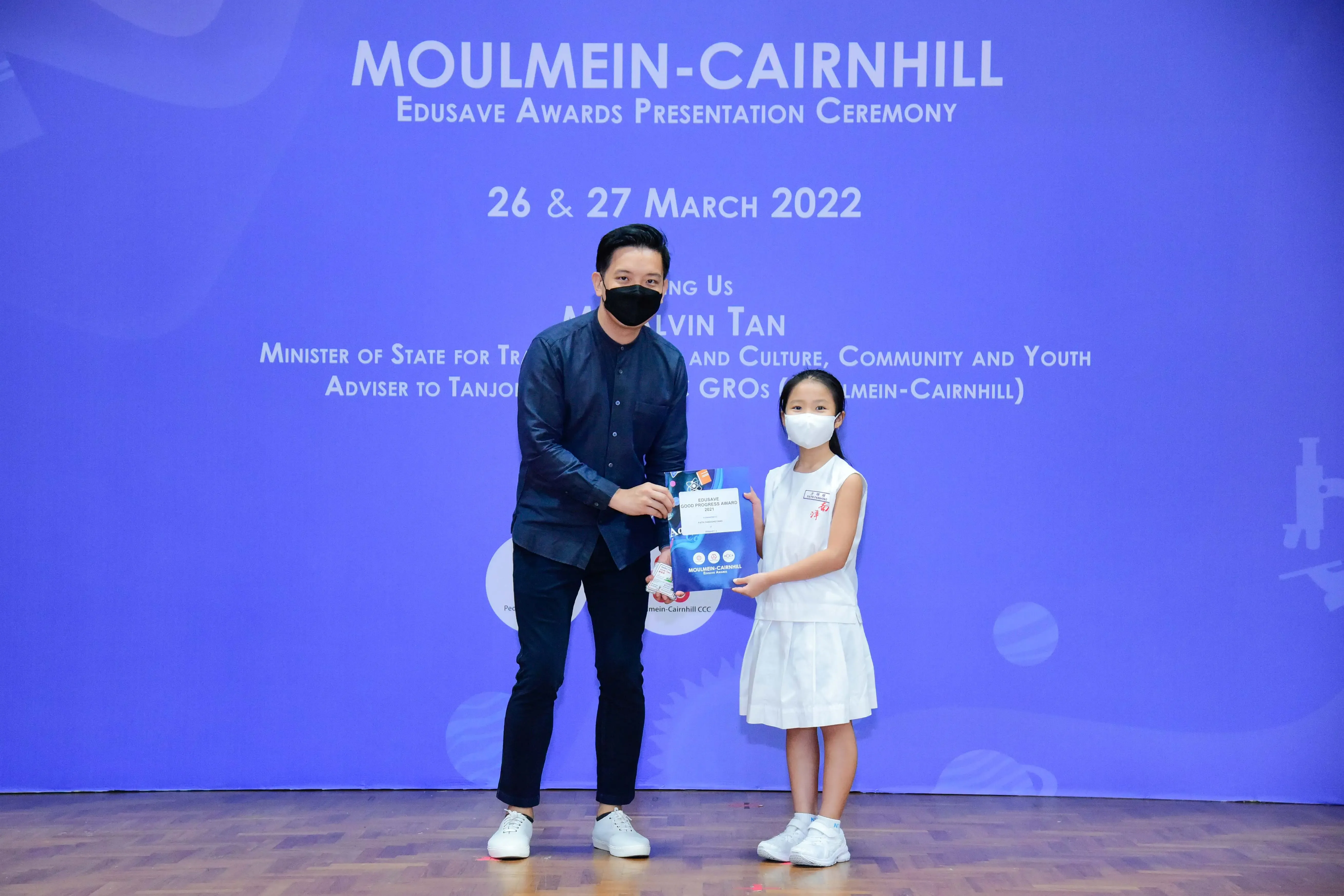 A masked man presents an award to a young girl in white at the Moulmein-Cairnhill Edusave Awards Ceremony, March 2022.