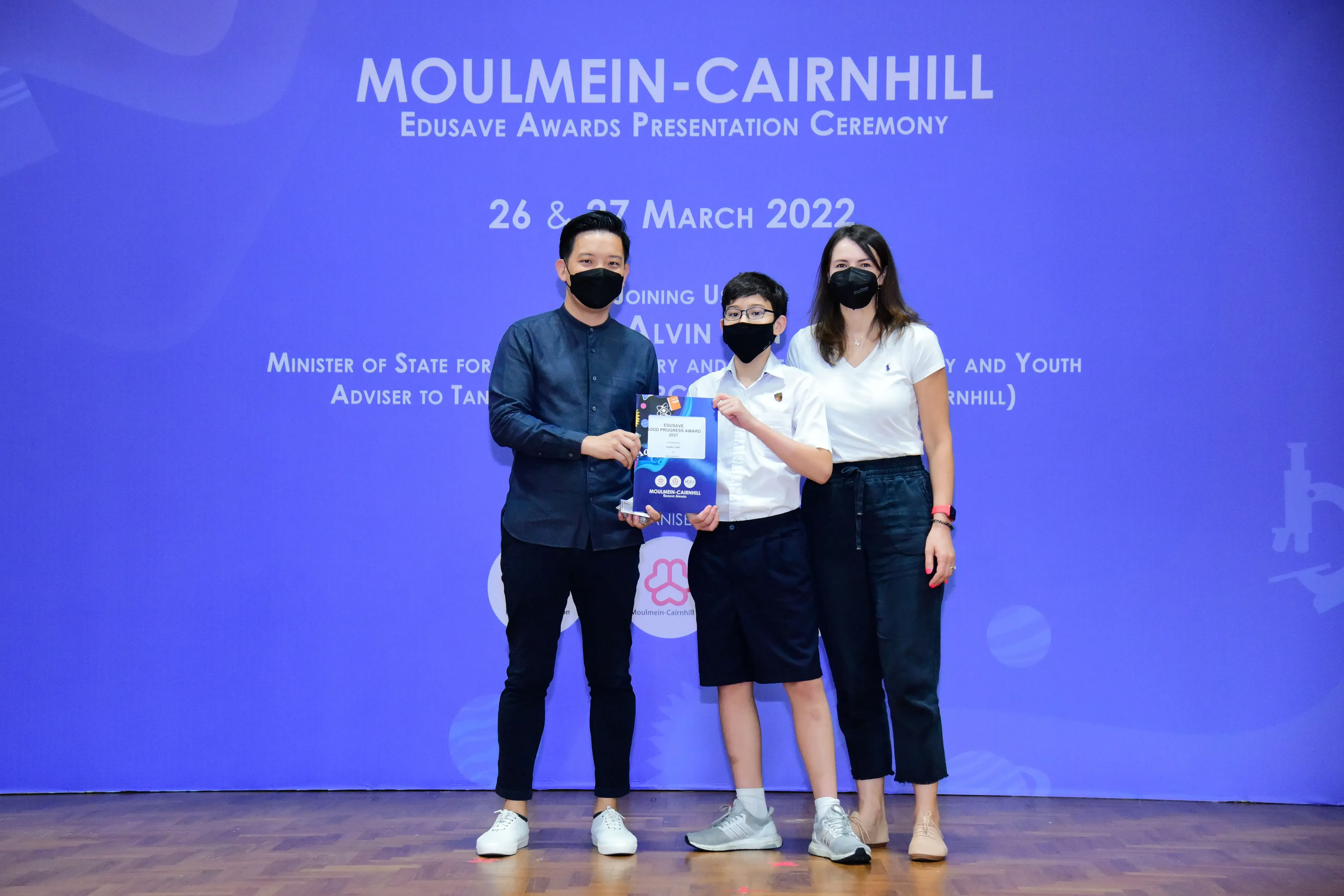 Three masked people hold an award at the Moulmein-Cairnhill Edusave Awards Presentation Ceremony, March 2022.