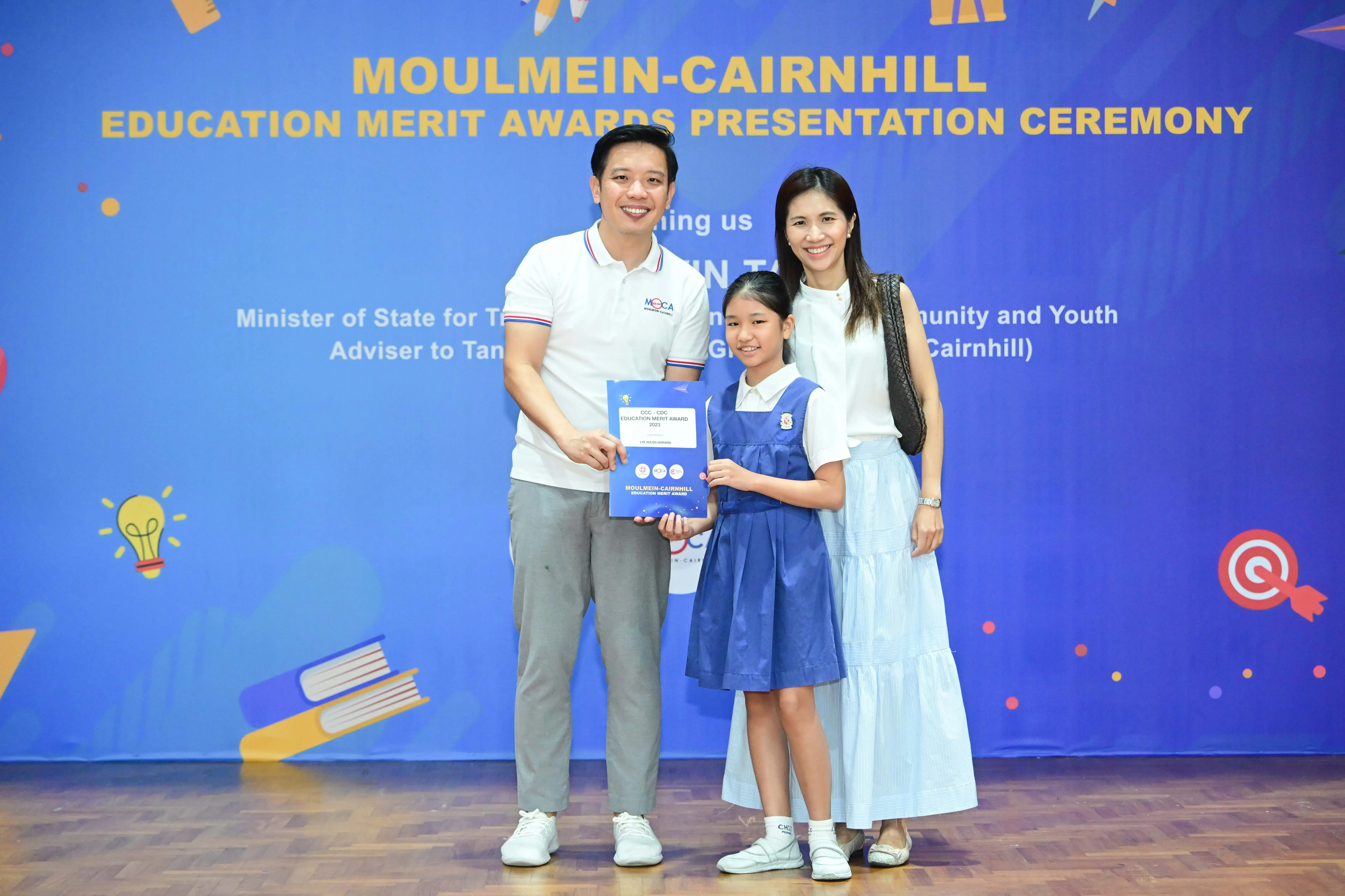 A girl in school uniform holds an award certificate with two adults at the Moulmein-Cairnhill Education Merit Awards ceremony.