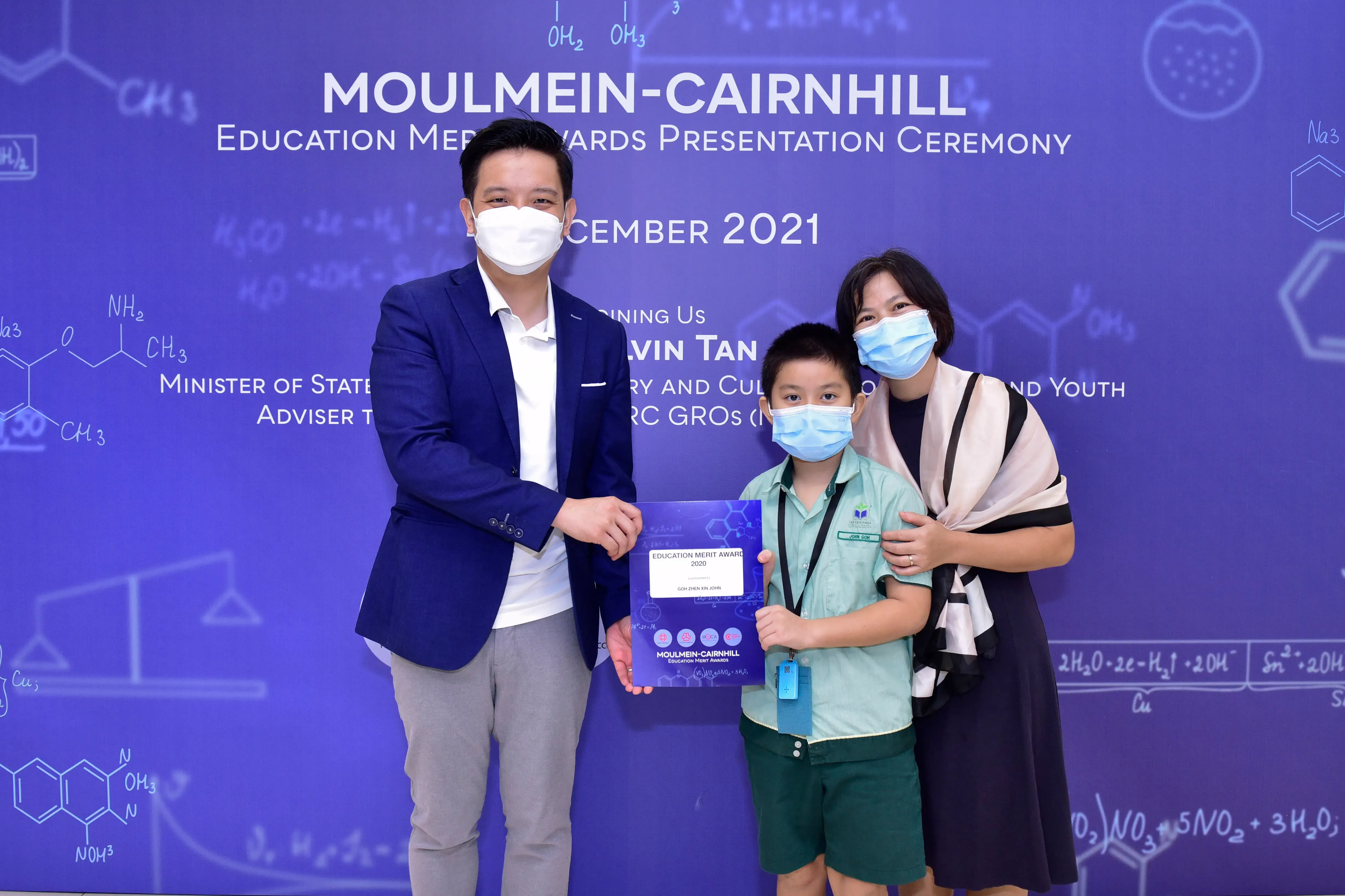 Three masked people pose at the Moulmein-Cairnhill Education Merit Awards Ceremony 2021, with a boy holding a certificate.