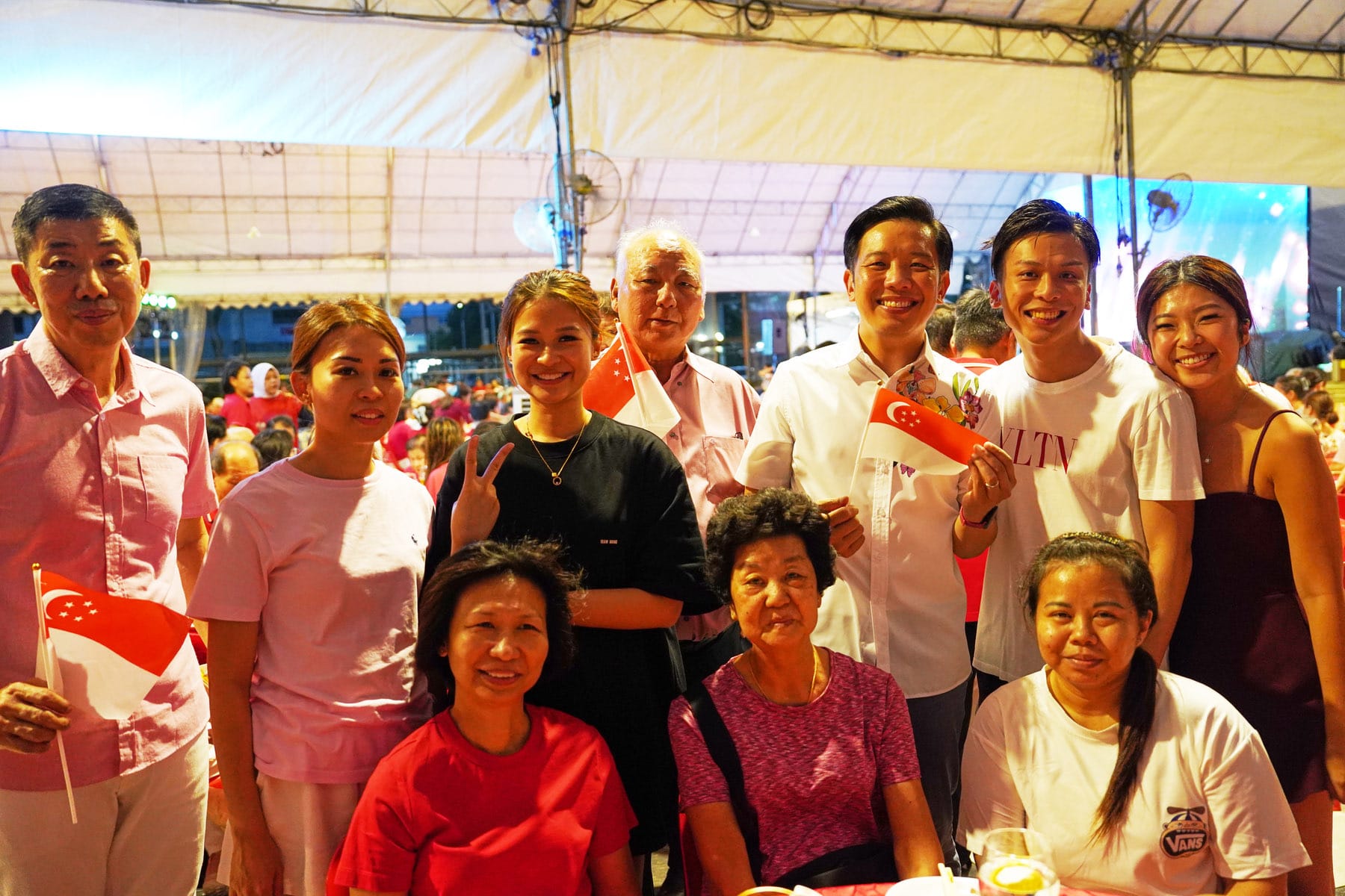 A group of smiling people posing together under a tent at a celebration, some holding small Singapore flags.