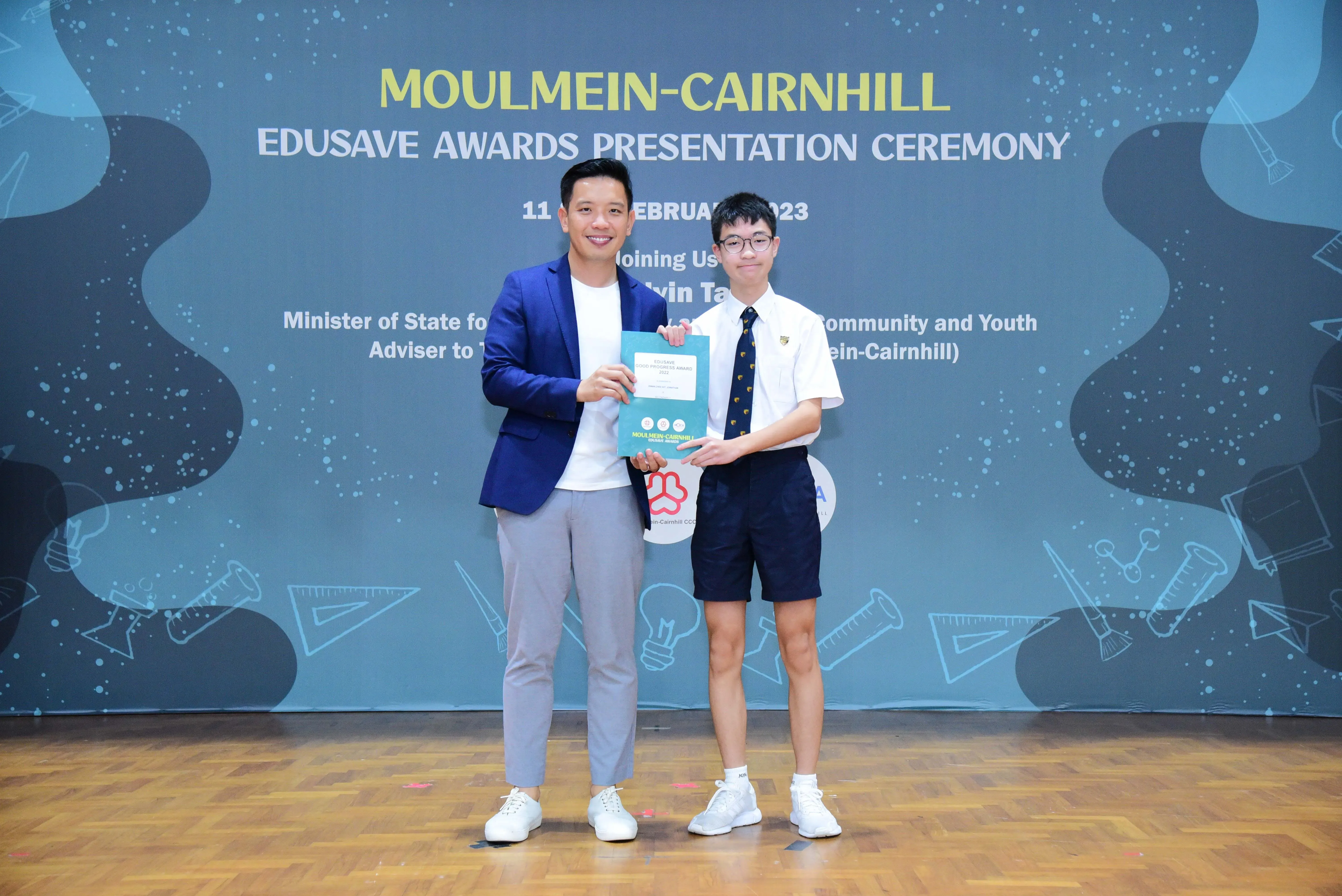 An adult in a blue blazer and a young student in uniform hold an award folder at the Moulmein-Cairnhill Edusave Ceremony.