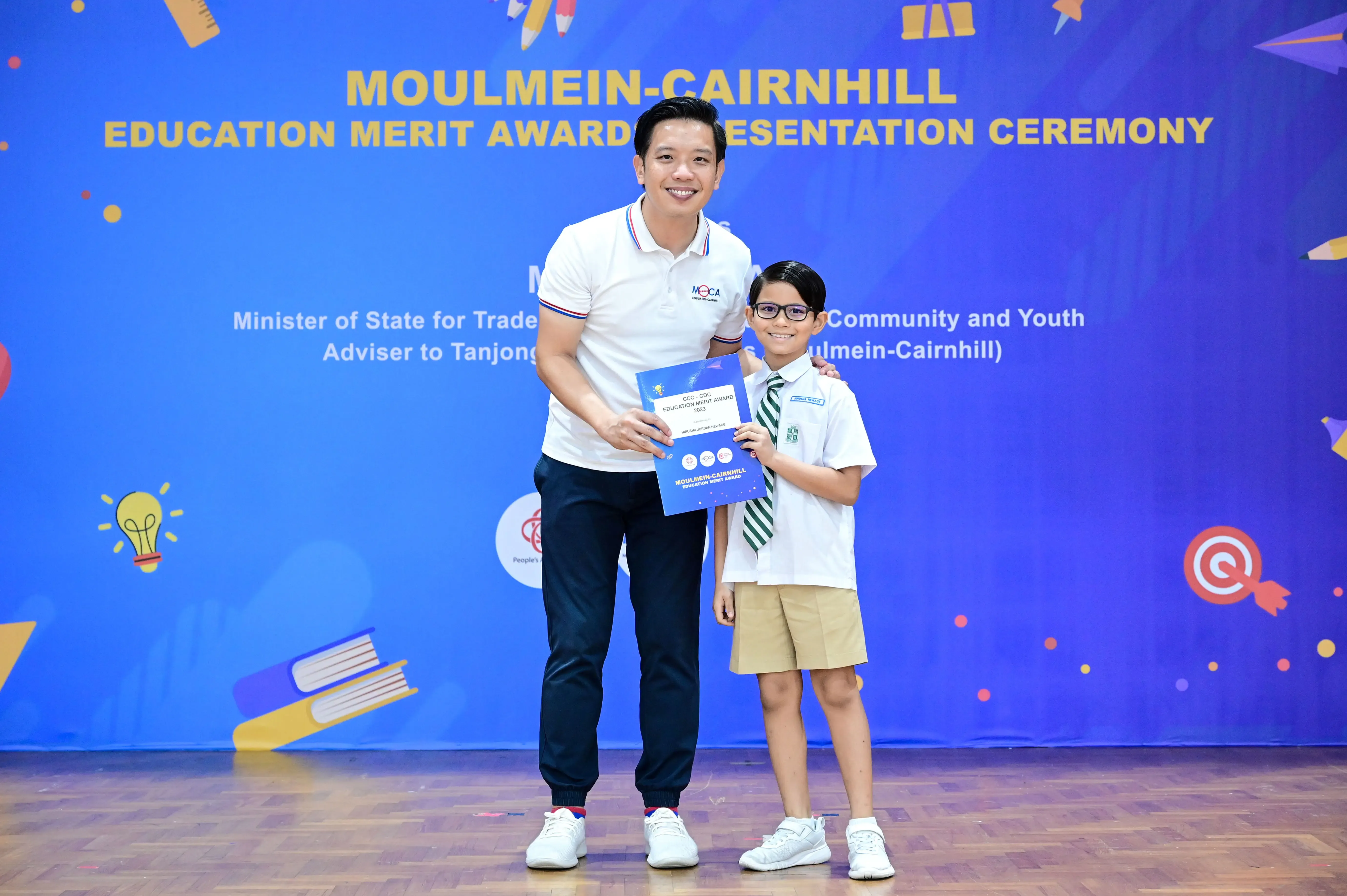 An adult and a uniformed boy hold a certificate at the Moulmein-Cairnhill Education Merit Award Presentation Ceremony.