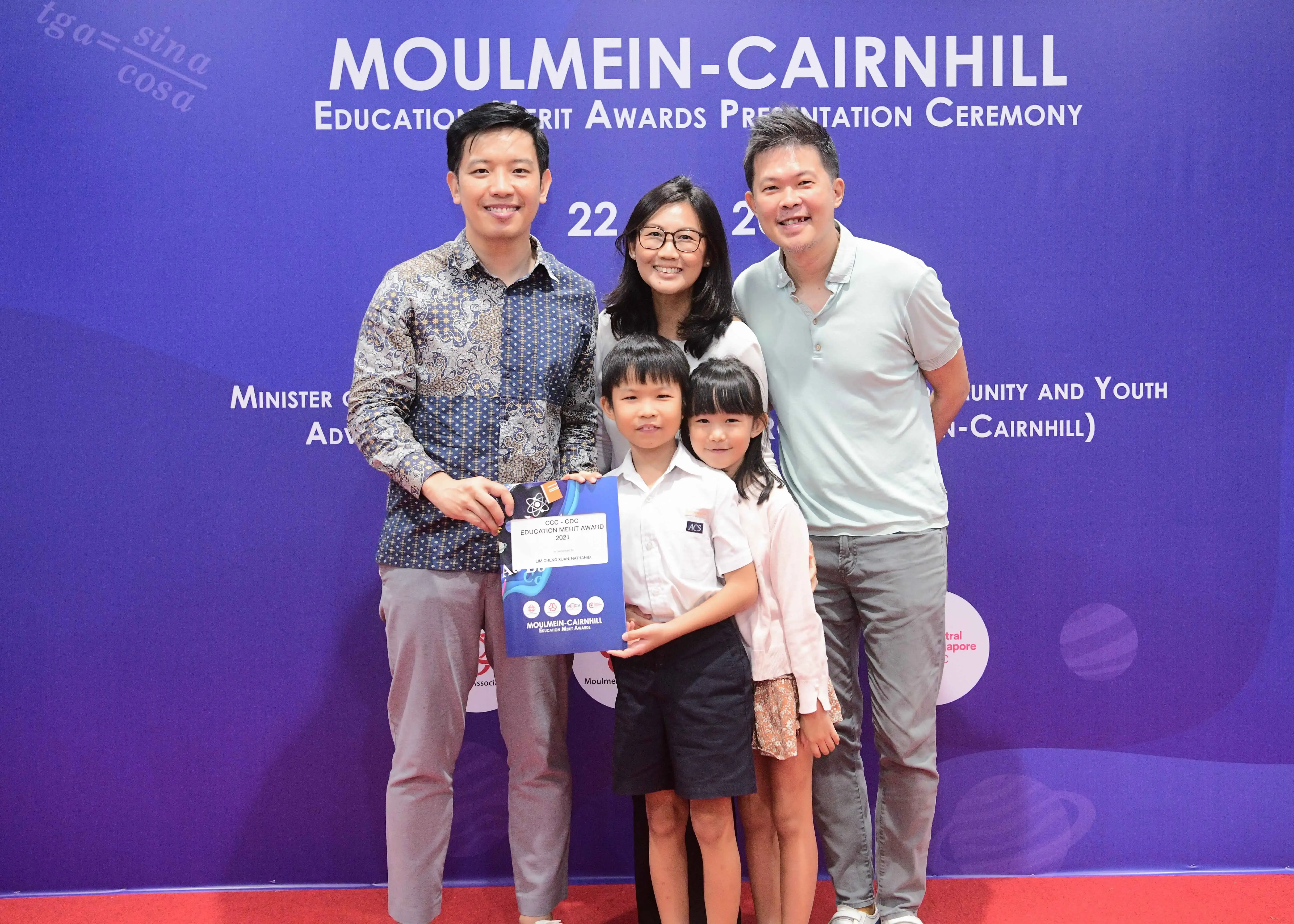 Five people pose smiling before a Moulmein-Cairnhill Education Merit Awards banner, holding an award certificate.