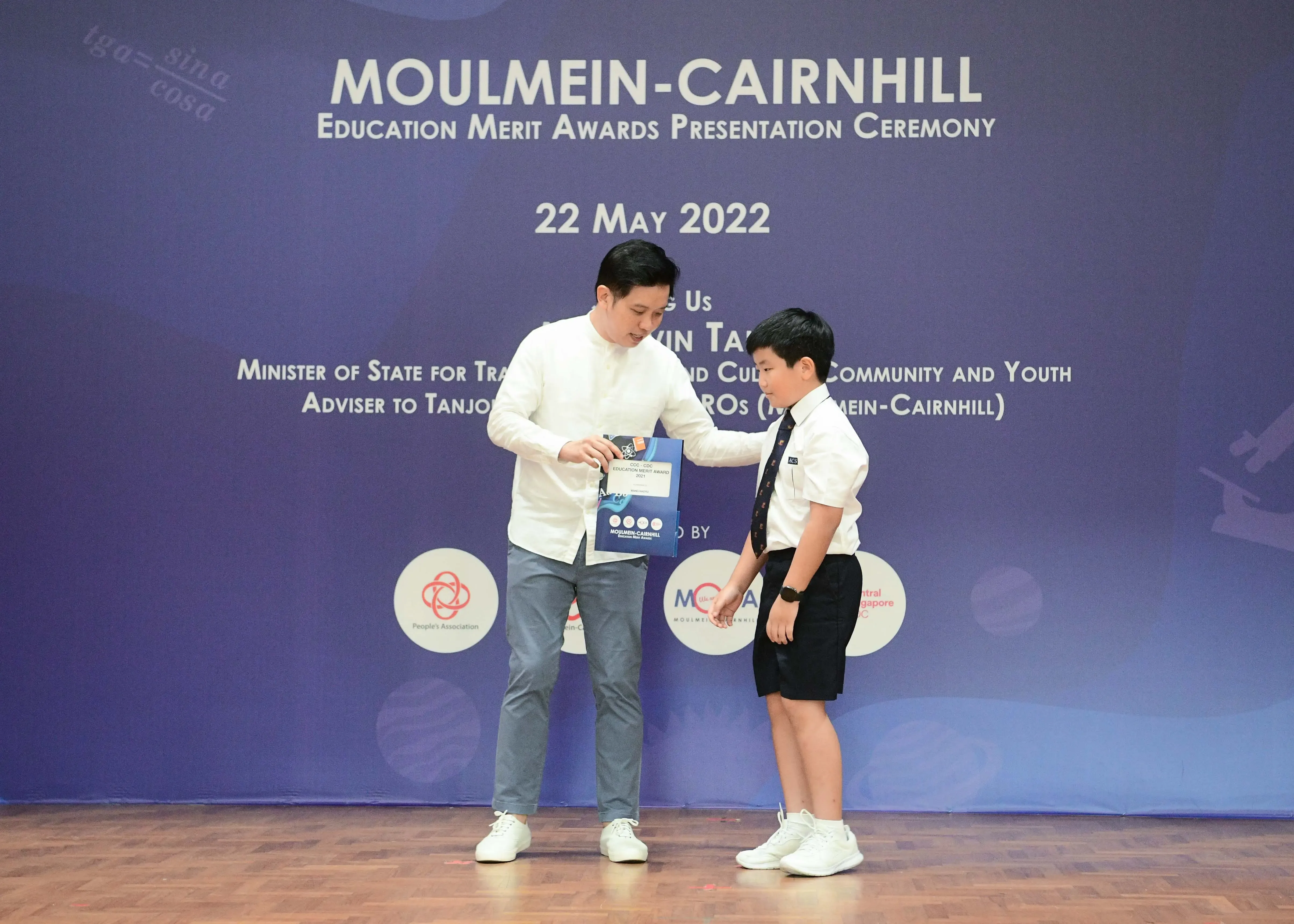 A man hands an award to a young boy in school uniform on stage at the Moulmein-Cairnhill Education Merit Awards, 22 May 2022.