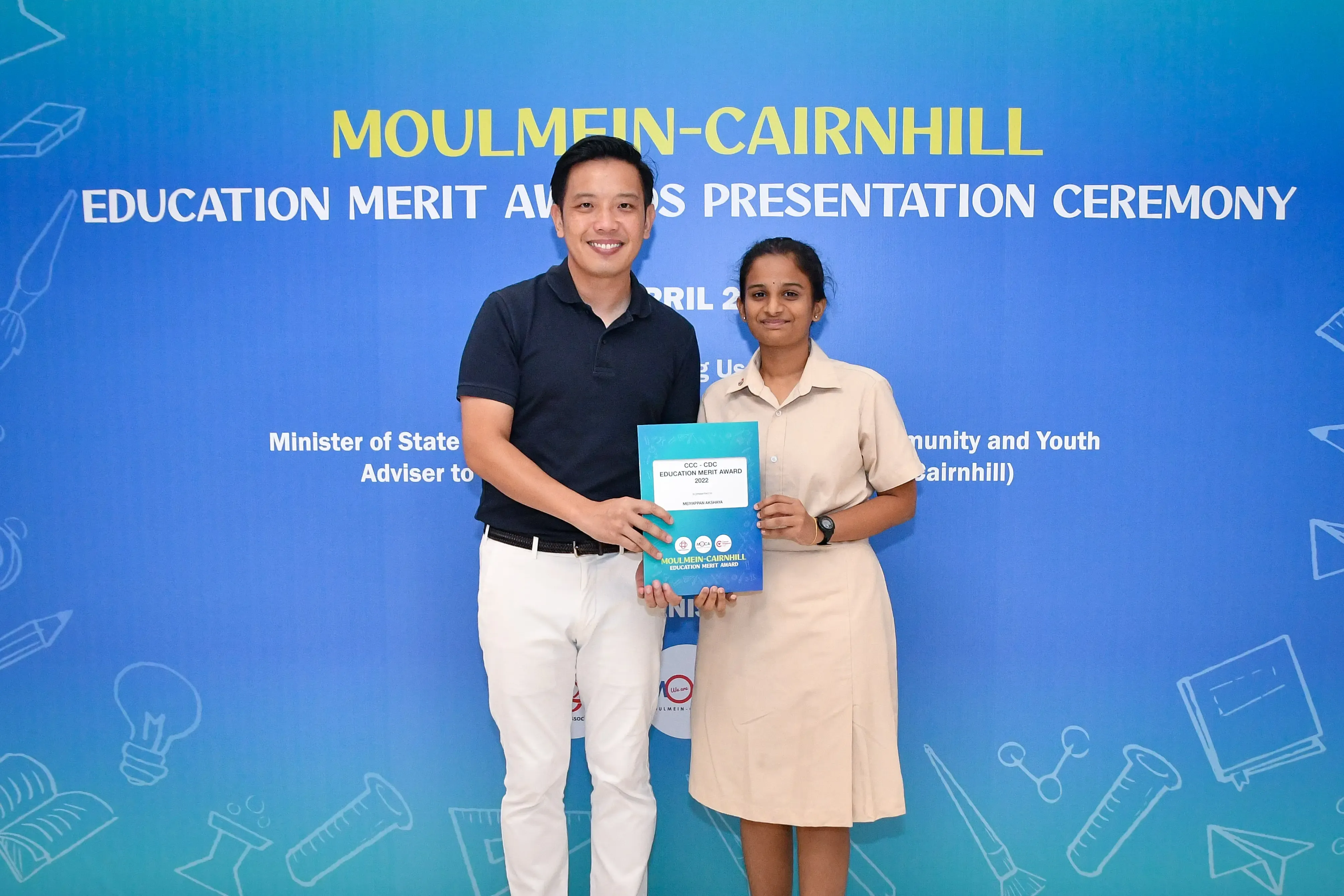 Two people hold a certificate together in front of a Moulmein-Cairnhill Education Merit Awards Presentation Ceremony banner.