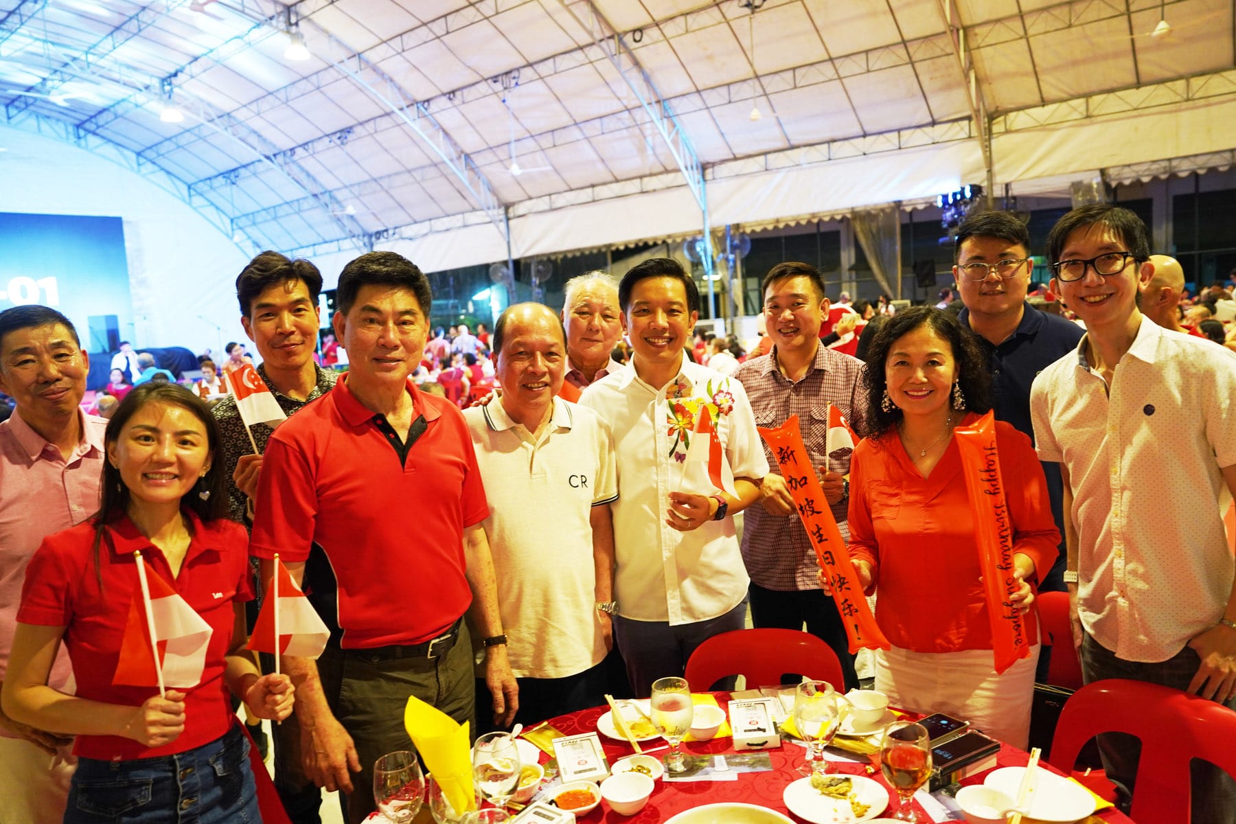 Group of people in red and white attire smiling at a Singapore National Day celebration under a tent.