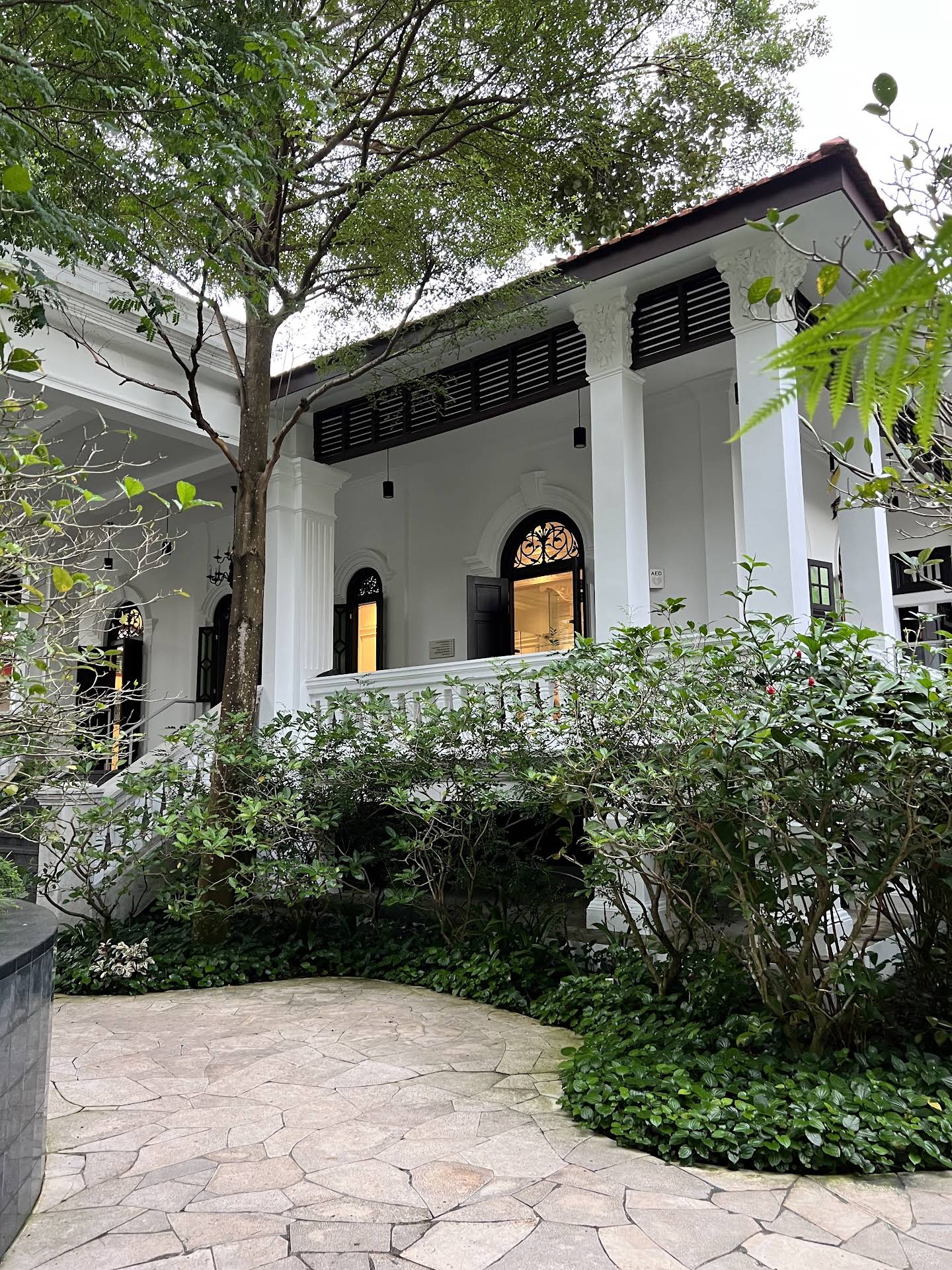 White colonial-style building with dark shutters and arched windows, surrounded by lush greenery and a stone pathway.