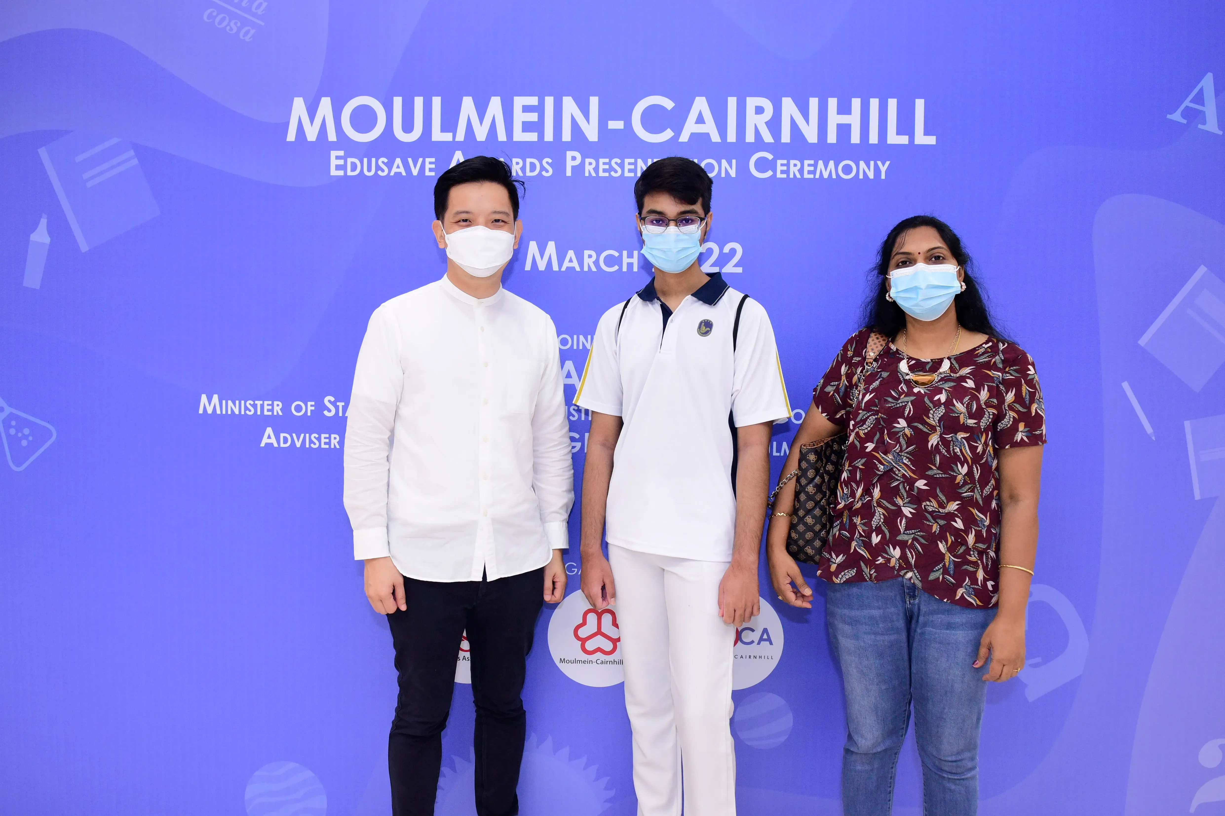 Three masked people pose before a blue Moulmein-Cairnhill Edusave Awards Presentation Ceremony backdrop dated March 2022.