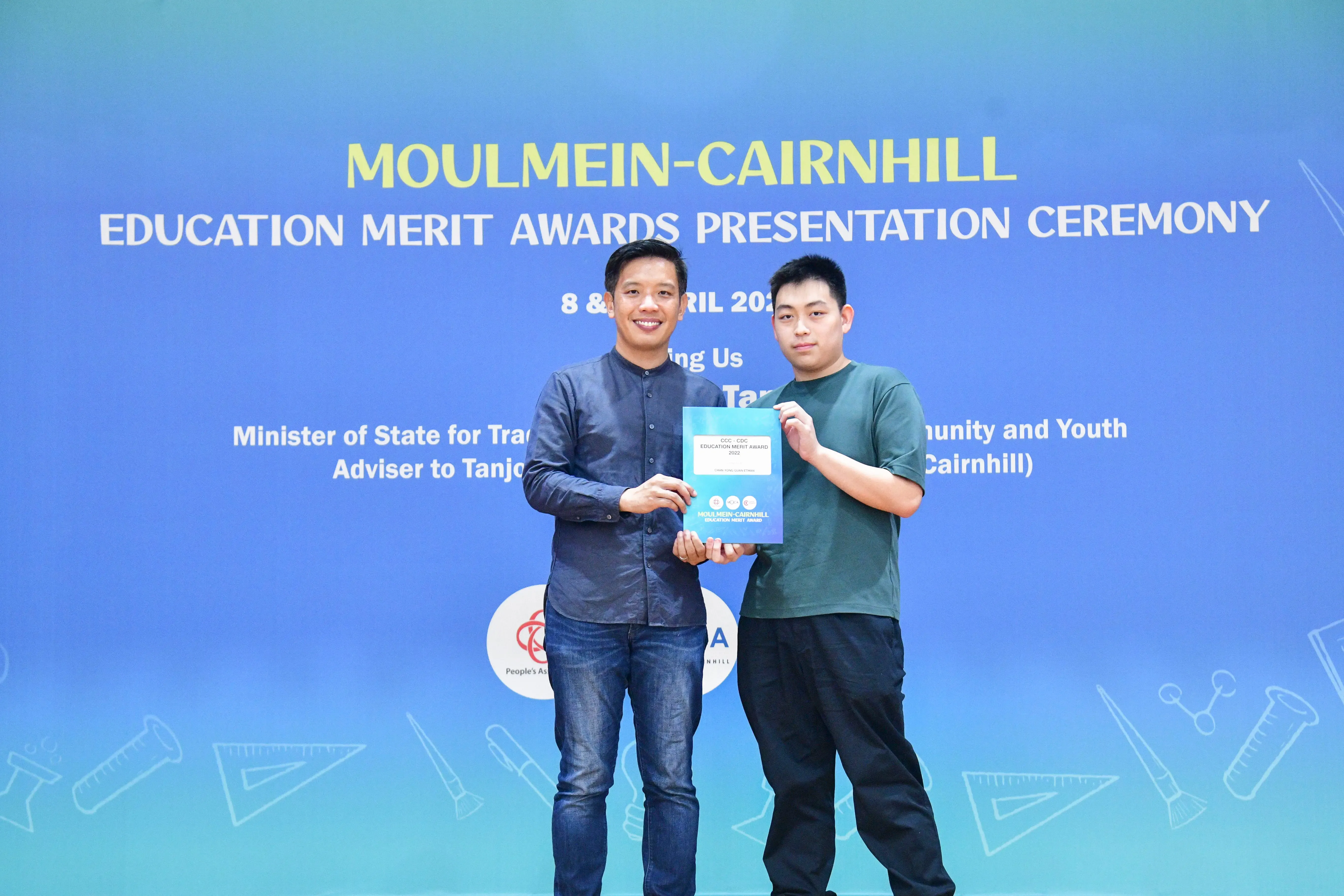 Two men hold a certificate together in front of a Moulmein-Cairnhill Education Merit Awards banner.