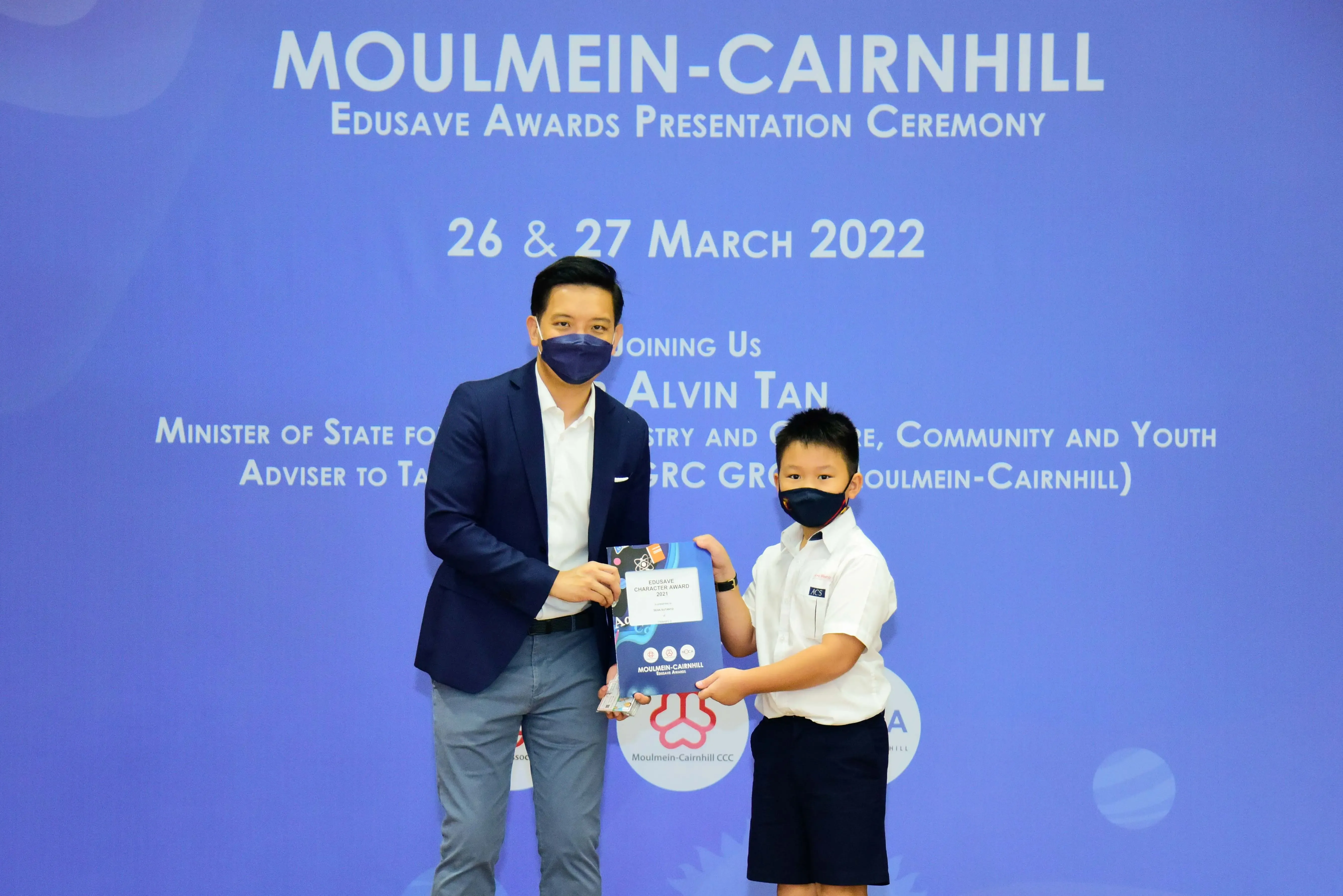 An adult and a young student in school uniform hold an award together at the Moulmein-Cairnhill Edusave Awards Ceremony 2022.