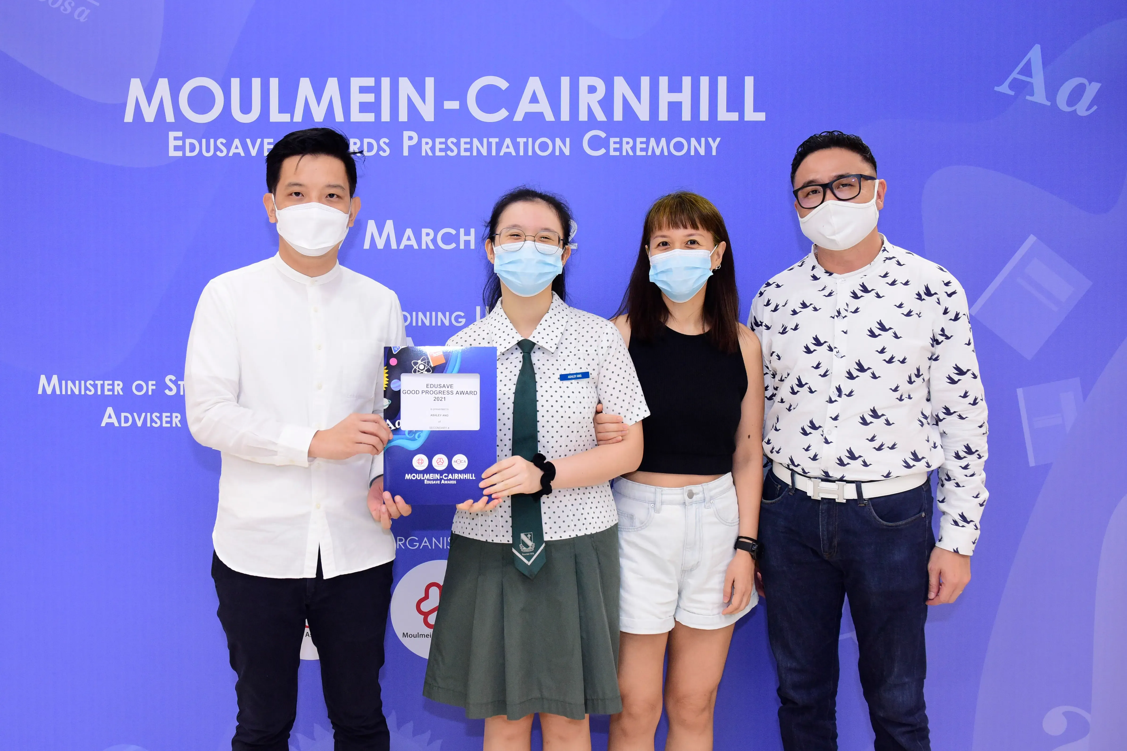 Four masked people pose at the Moulmein-Cairnhill Edusave Awards Ceremony, with a student holding a certificate.