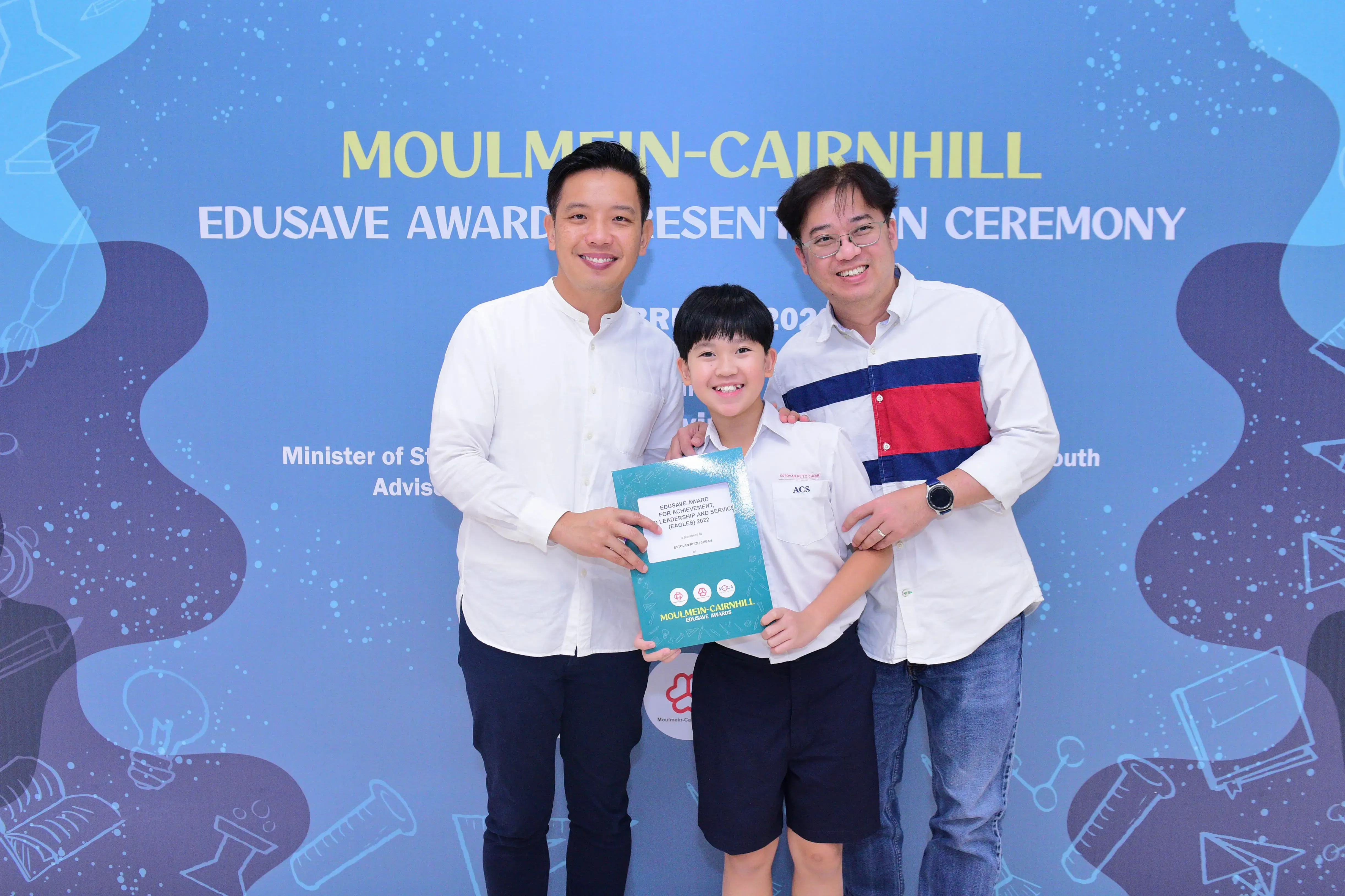 Three smiling people pose at the Moulmein-Cairnhill Edusave Award Presentation Ceremony, with a child holding a certificate.