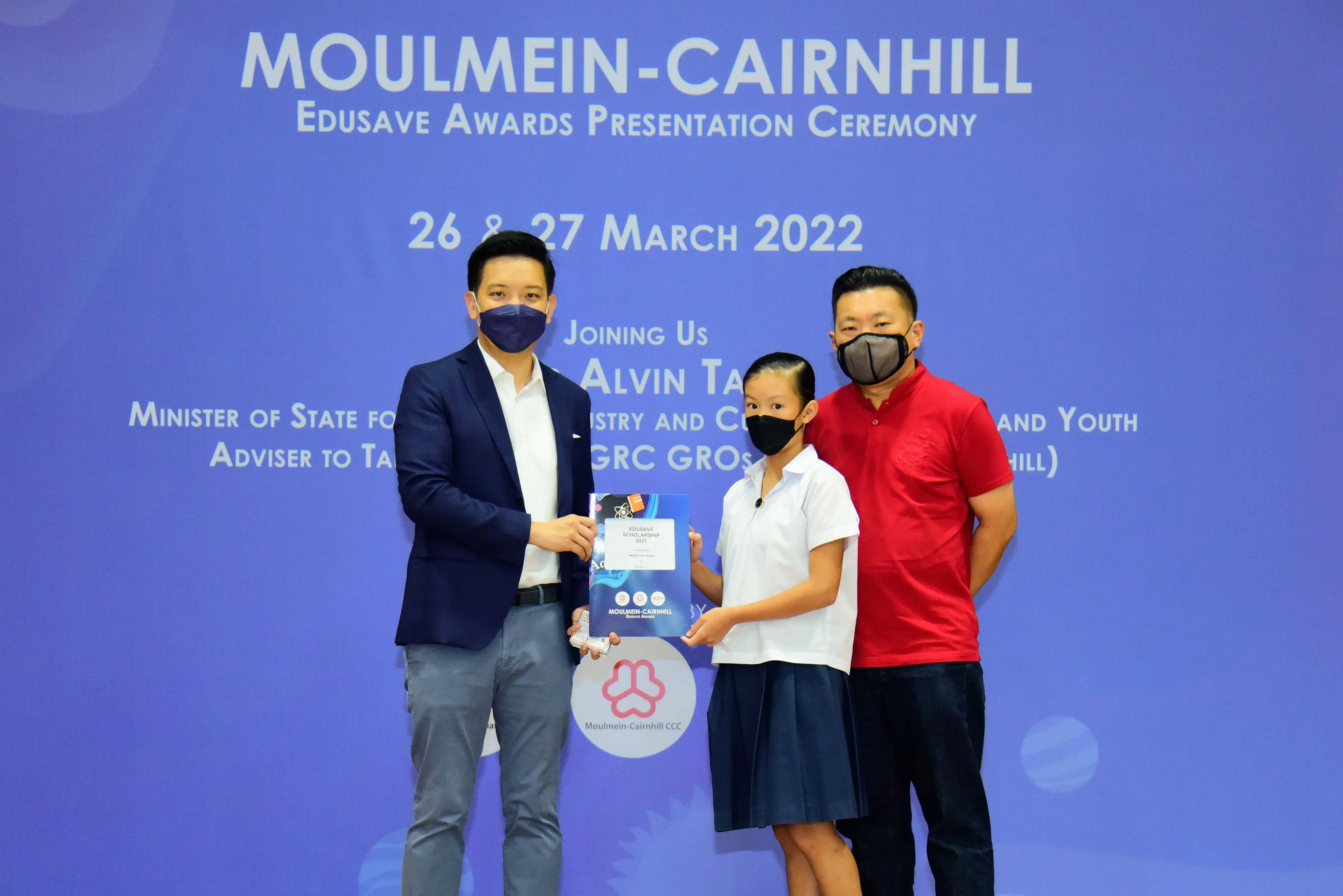 Two masked adults and a masked child holding an award at the Moulmein-Cairnhill Edusave Awards Ceremony, March 2022.