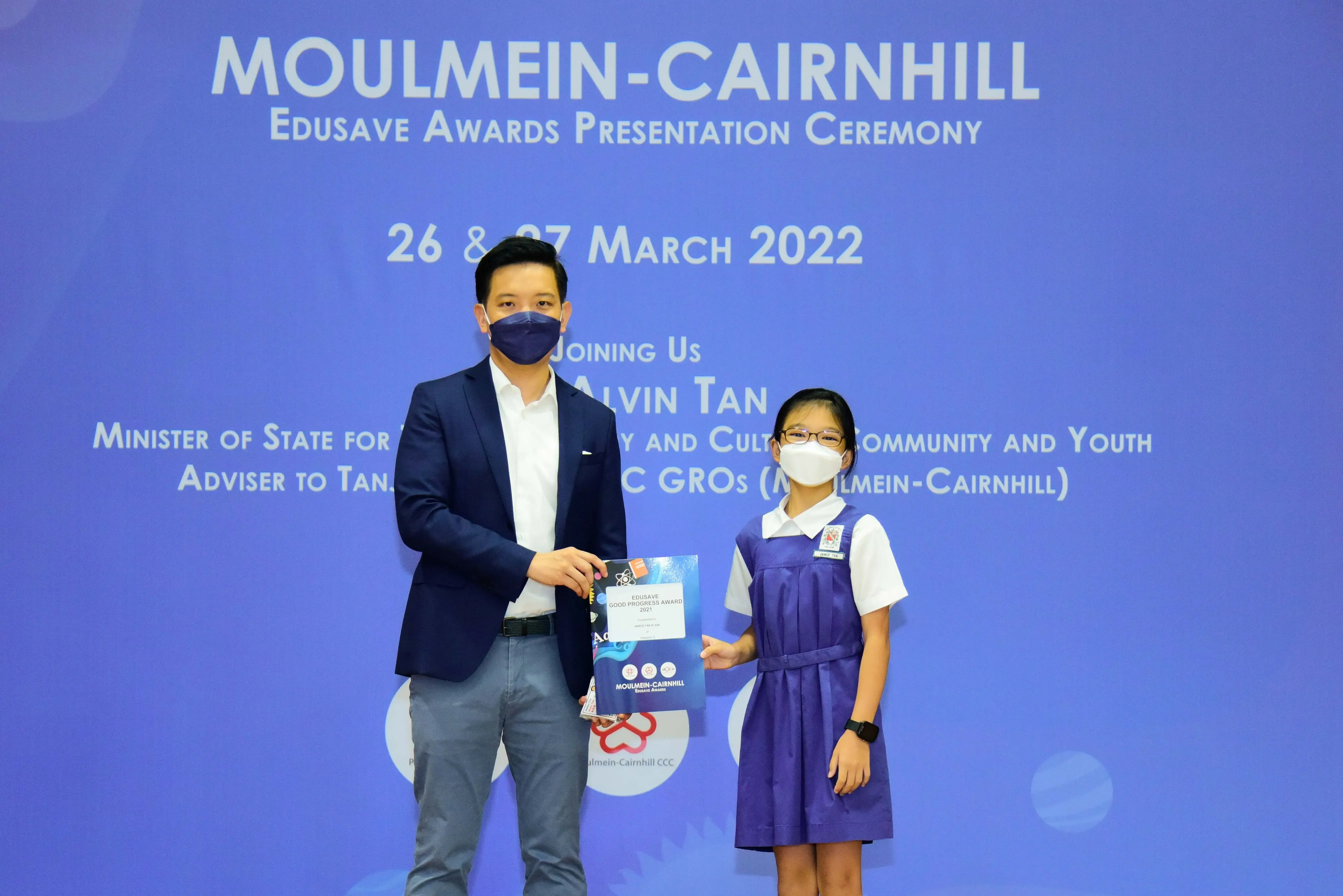 A masked man in a suit presents an award certificate to a masked young girl in school uniform at a 2022 ceremony.