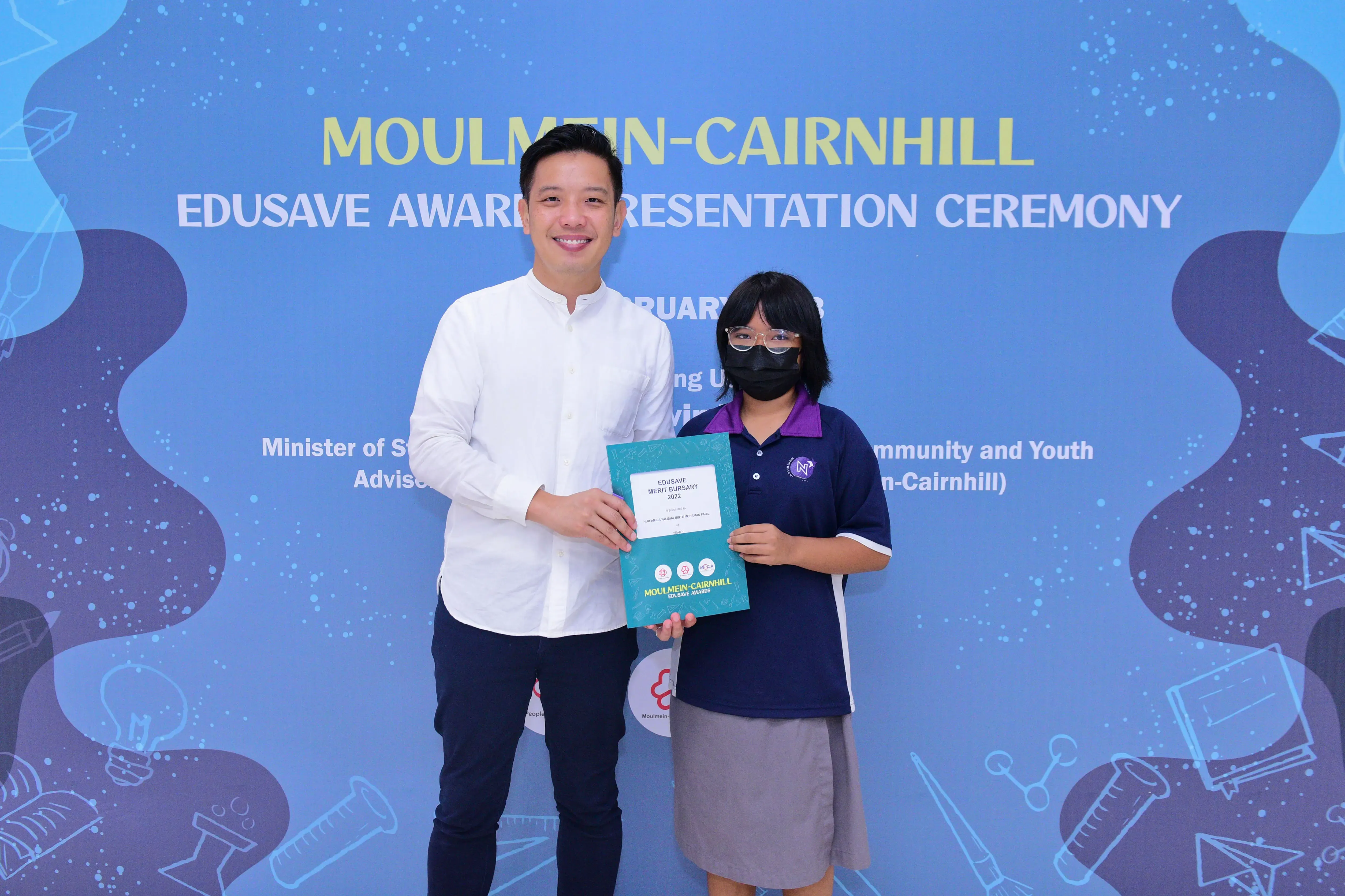 A man and a masked student hold a Moulmein-Cairnhill Edusave Merit Bursary certificate at an awards ceremony.
