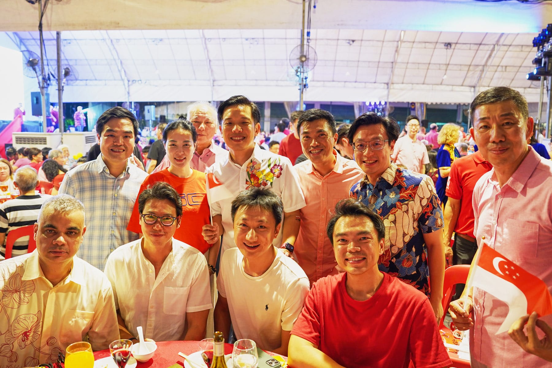 A group of smiling people pose together at an outdoor event, some holding small Singapore flags, under a large tent.