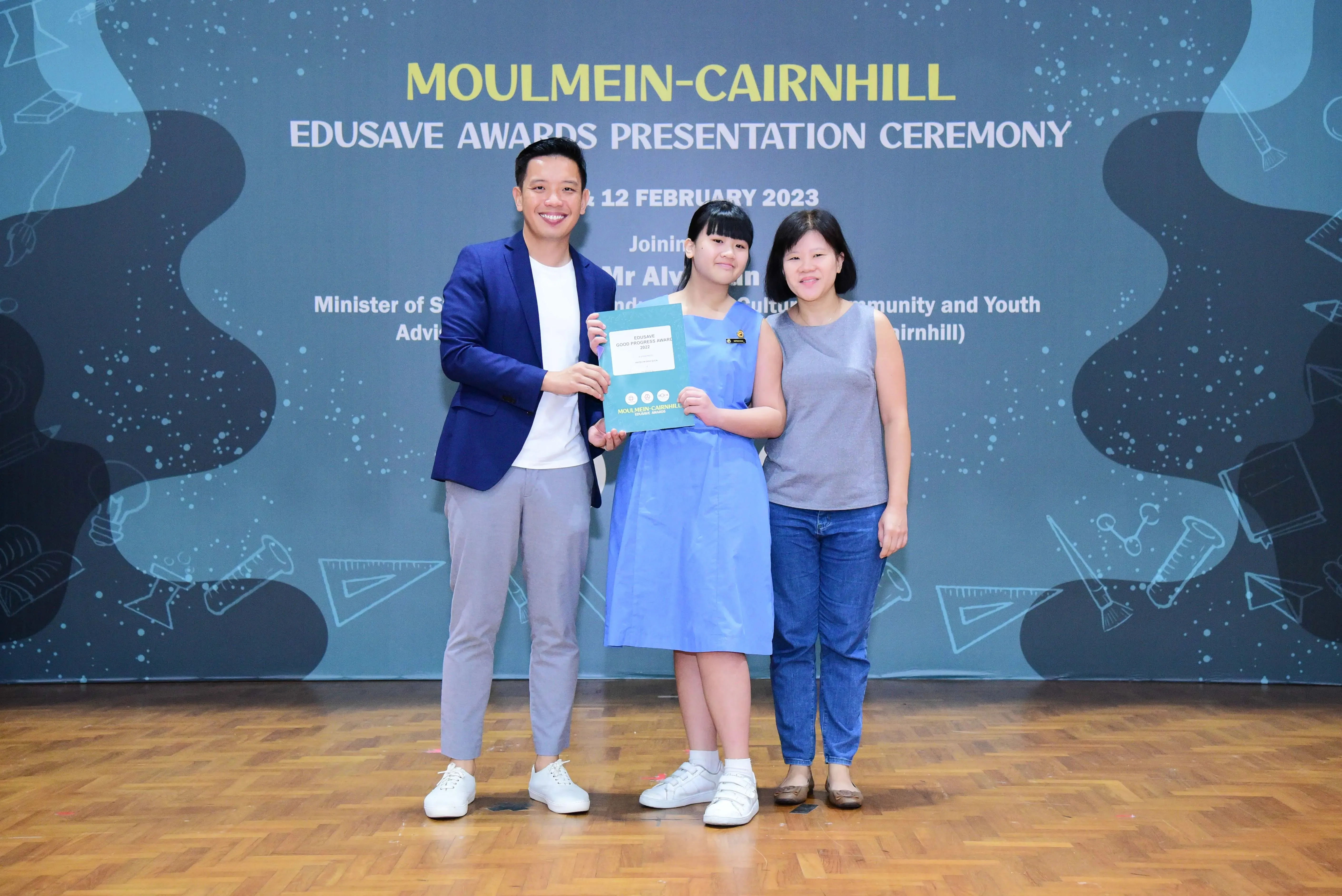 Three people pose at the Moulmein-Cairnhill Edusave Awards Presentation Ceremony, with a girl holding a certificate.