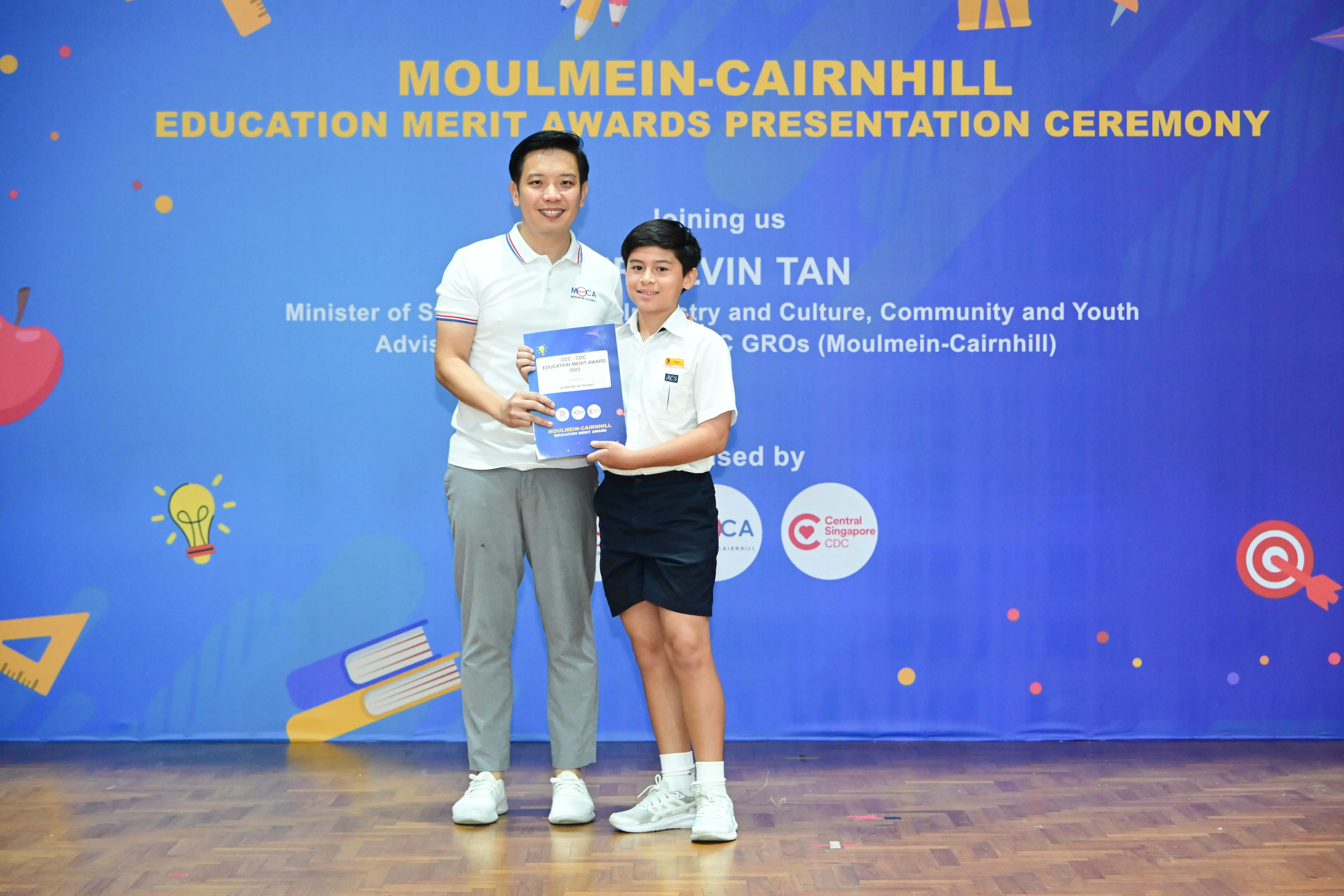 An adult and a child hold a certificate together in front of a Moulmein-Cairnhill Education Merit Awards banner.