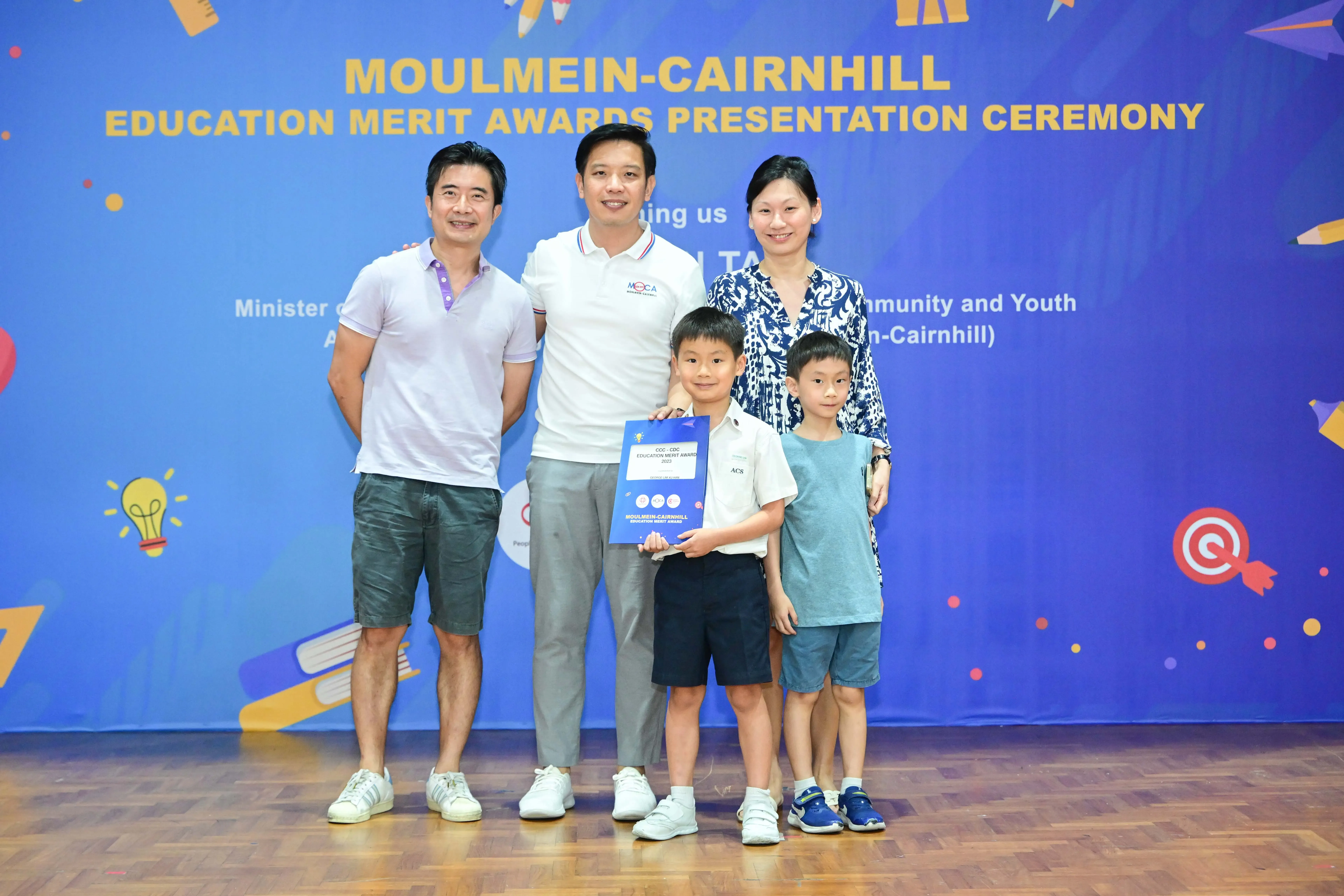 Three adults and two boys pose before a Moulmein-Cairnhill Education Merit Awards Presentation Ceremony banner.
