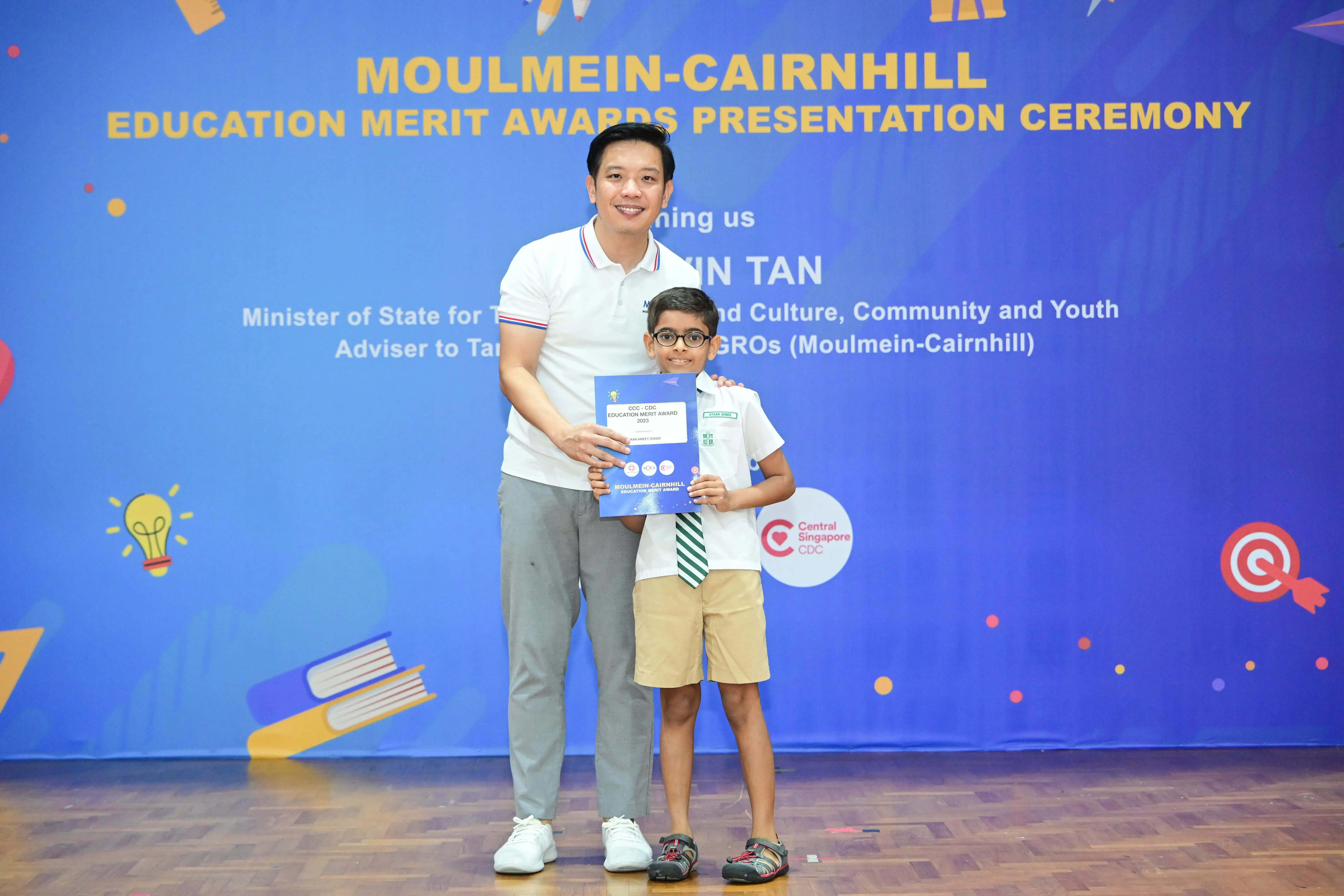 A man and a young boy in school uniform hold a certificate at the Moulmein-Cairnhill Education Merit Awards ceremony.
