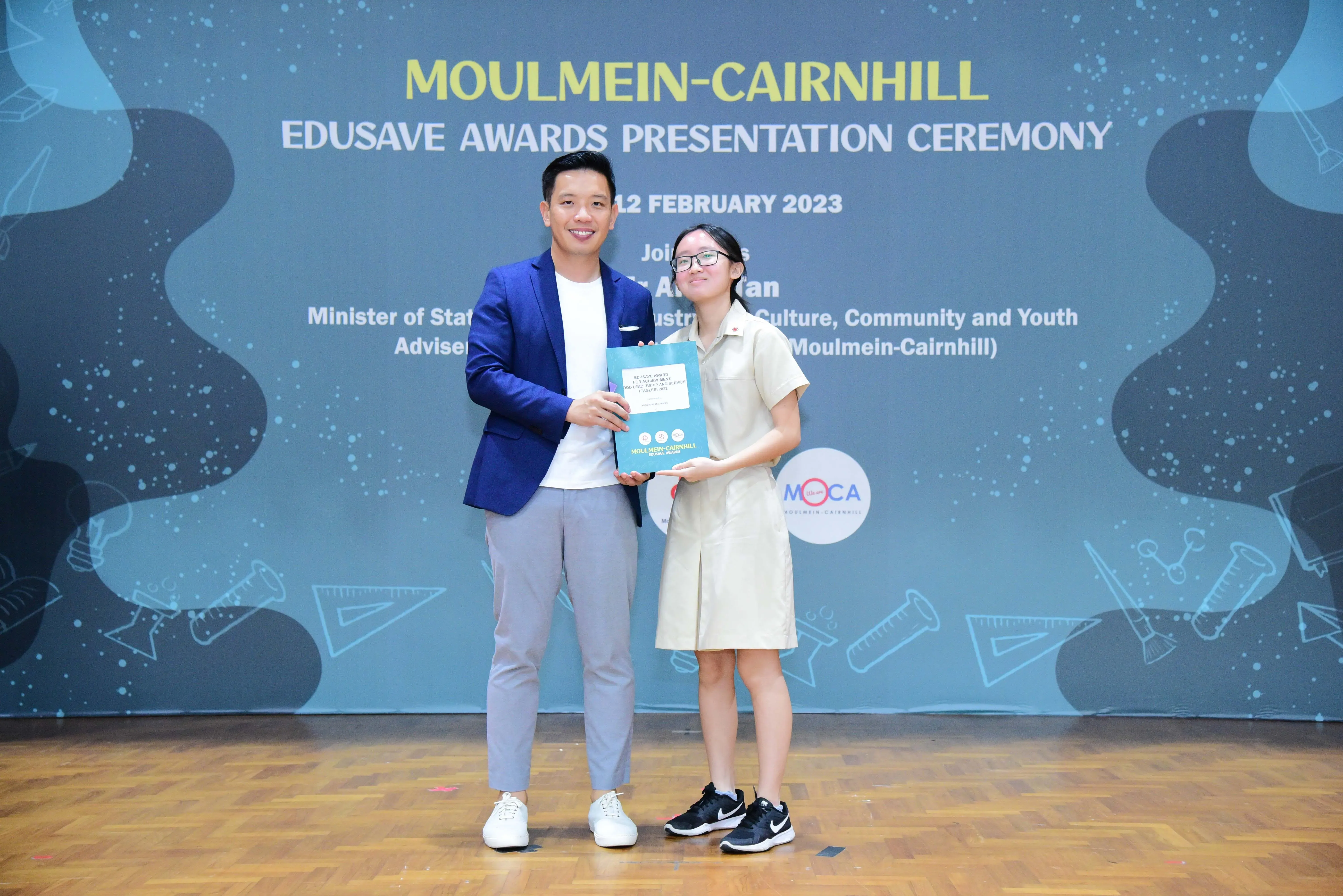 A man in a blue blazer and a young girl in school uniform hold a certificate at the Moulmein-Cairnhill Edusave Awards 2023.