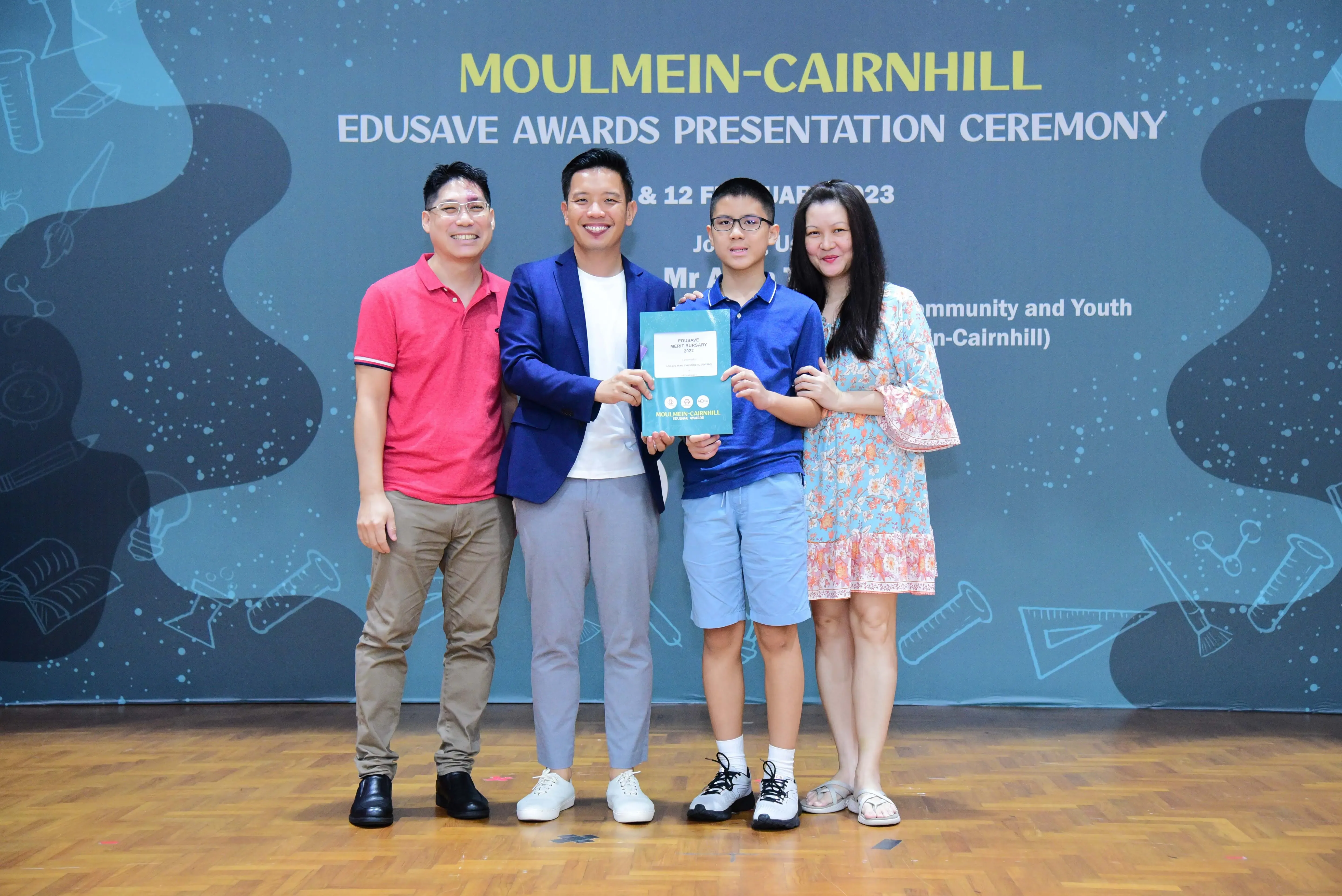 Four people pose on a stage at the Moulmein-Cairnhill Edusave Awards Presentation Ceremony, holding a certificate.