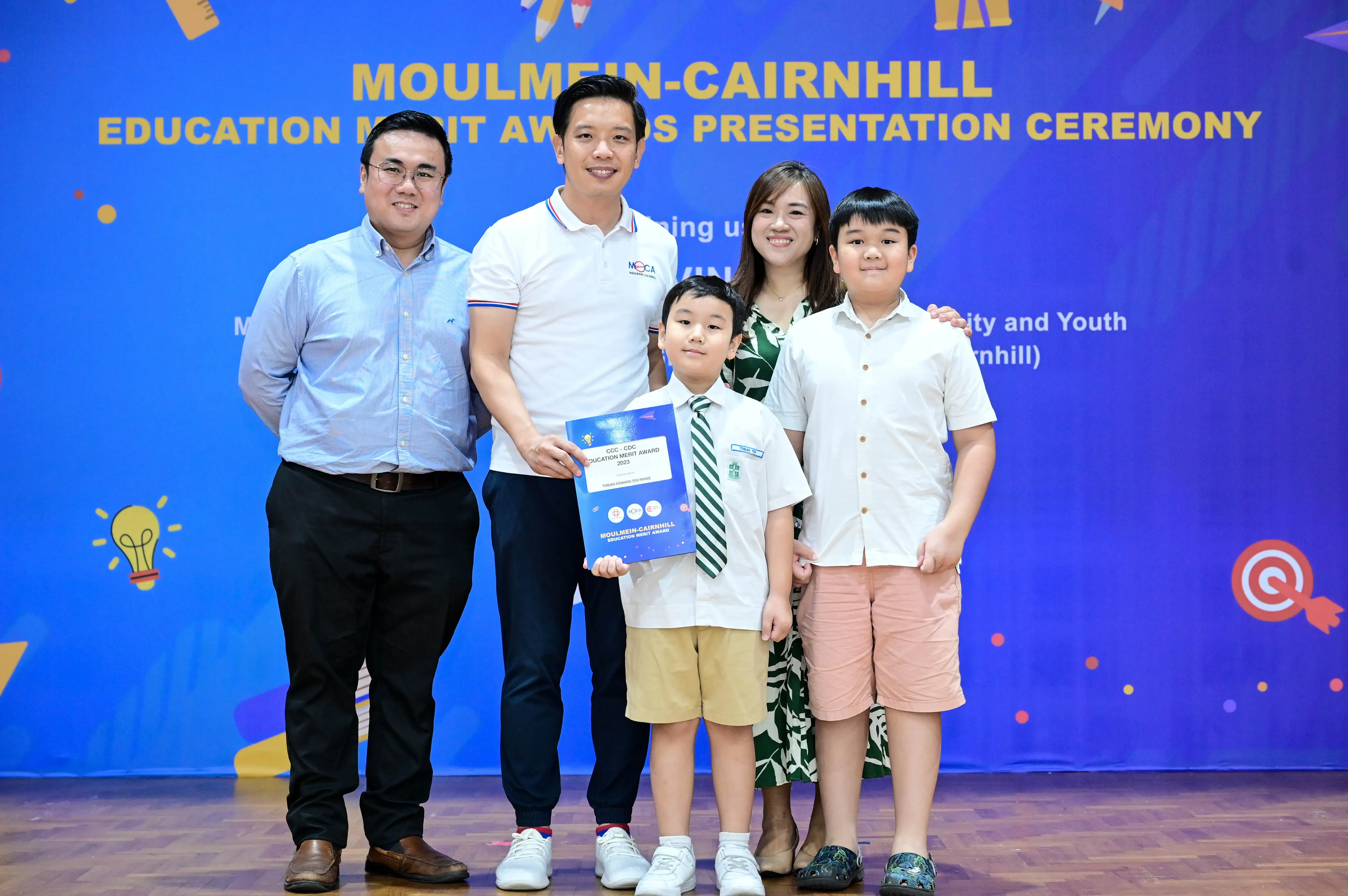 Five people pose at the Moulmein-Cairnhill Education Merit Awards Presentation Ceremony, with a boy holding a certificate.