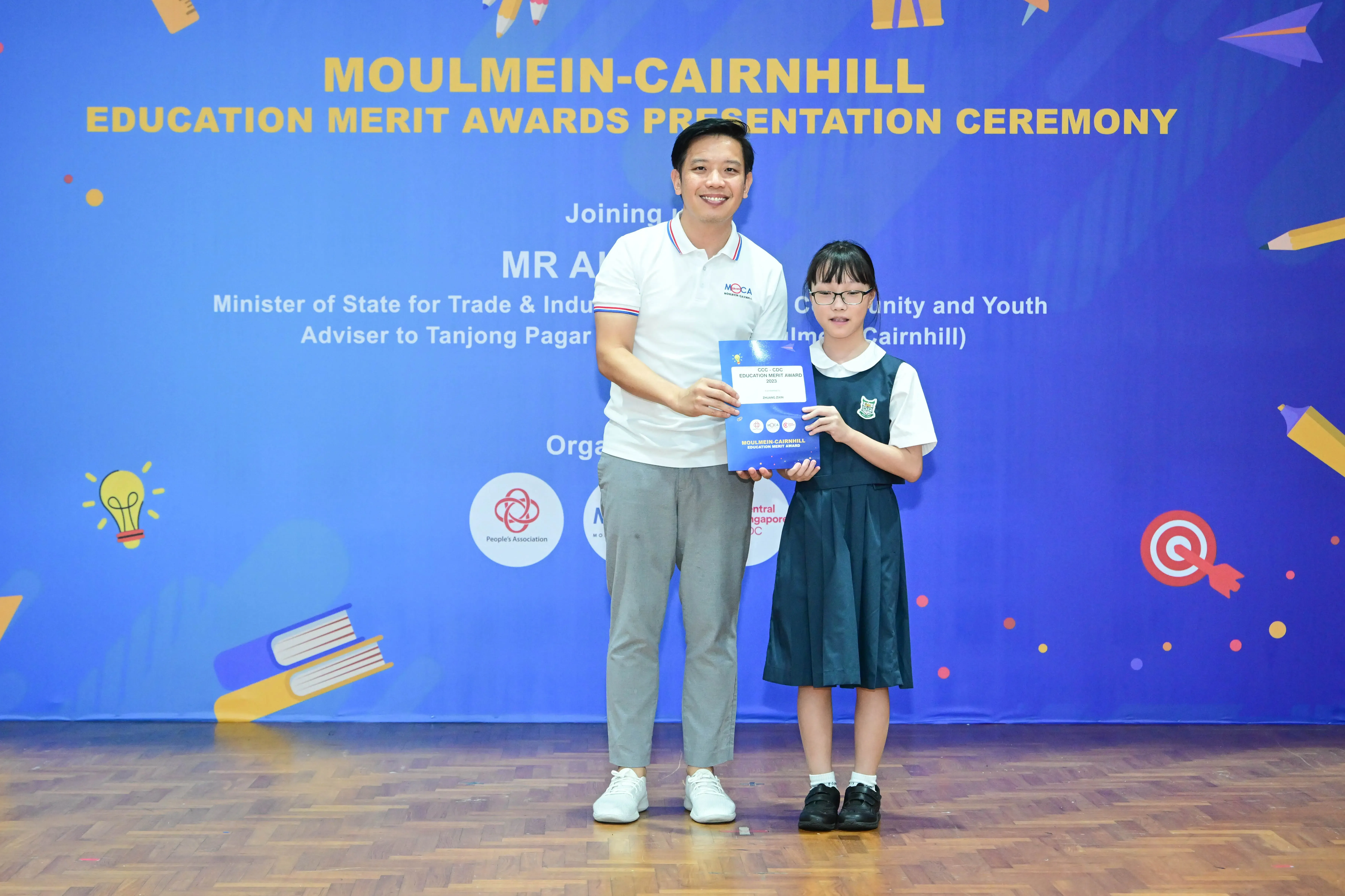 Man and young girl in school uniform hold an award certificate at Moulmein-Cairnhill Education Merit Awards ceremony.