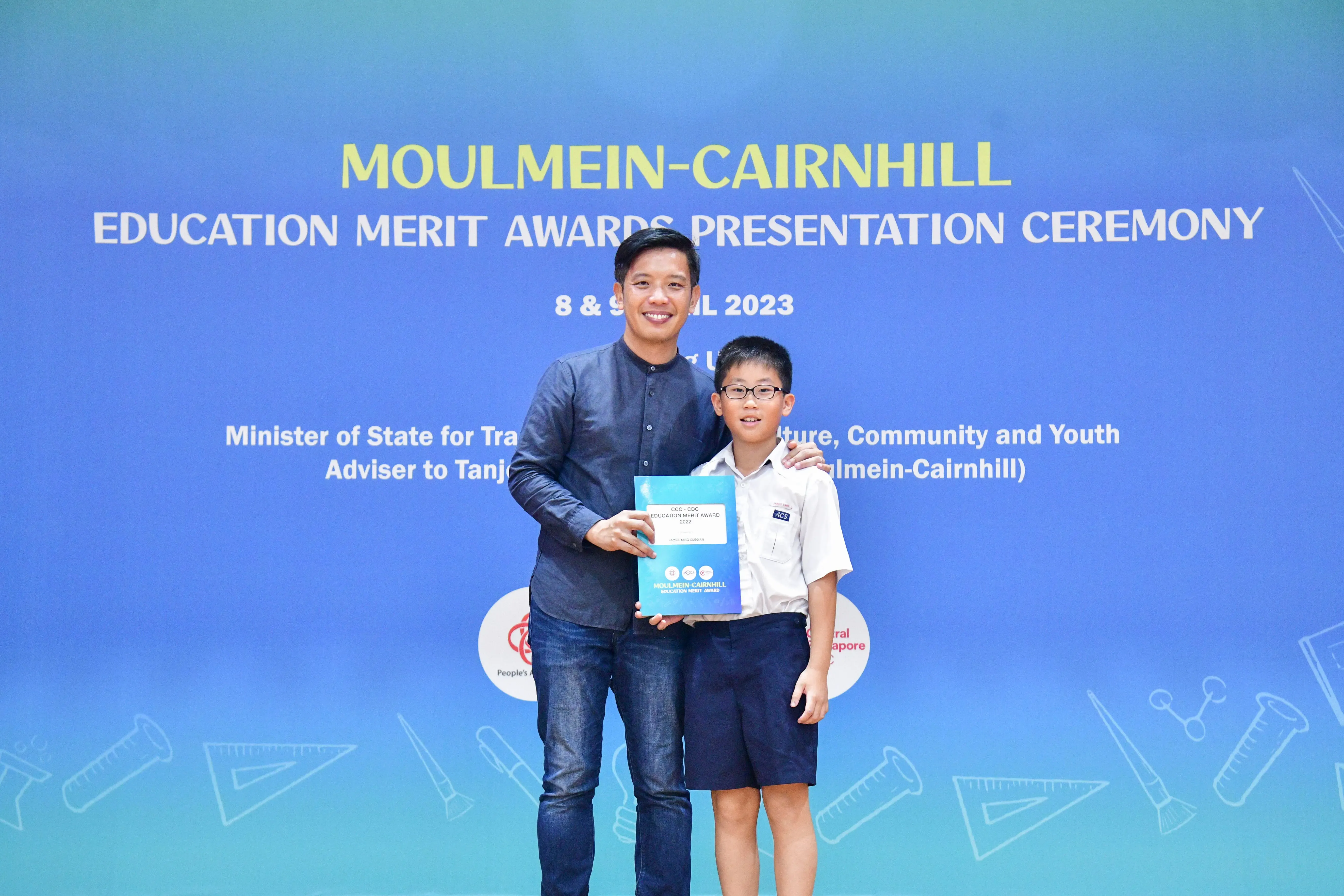 An adult man and a young boy hold a certificate in front of a Moulmein-Cairnhill Education Merit Awards 2023 banner.