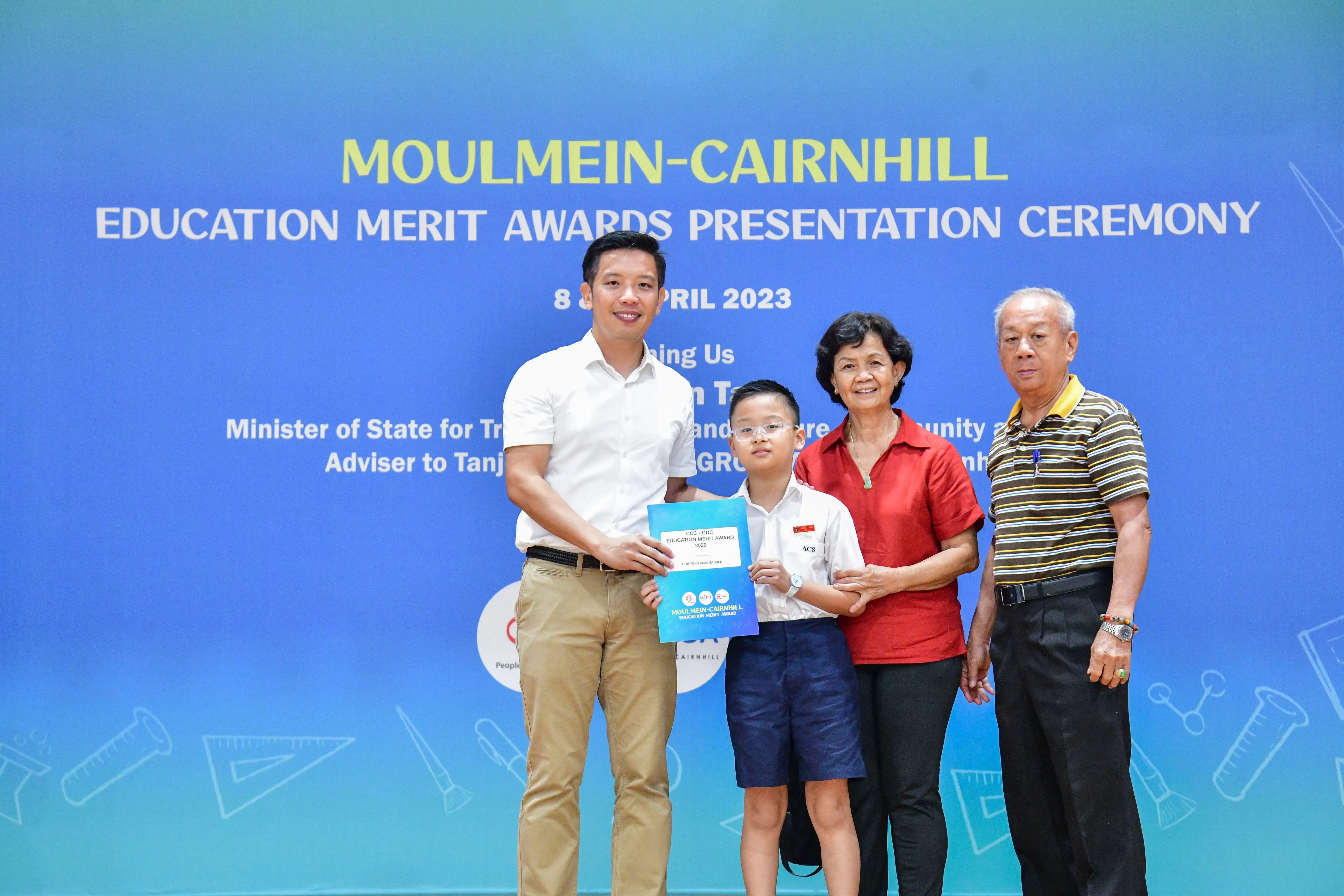 A young boy holds an award certificate with three adults at the Moulmein-Cairnhill Education Merit Awards Ceremony 2023.