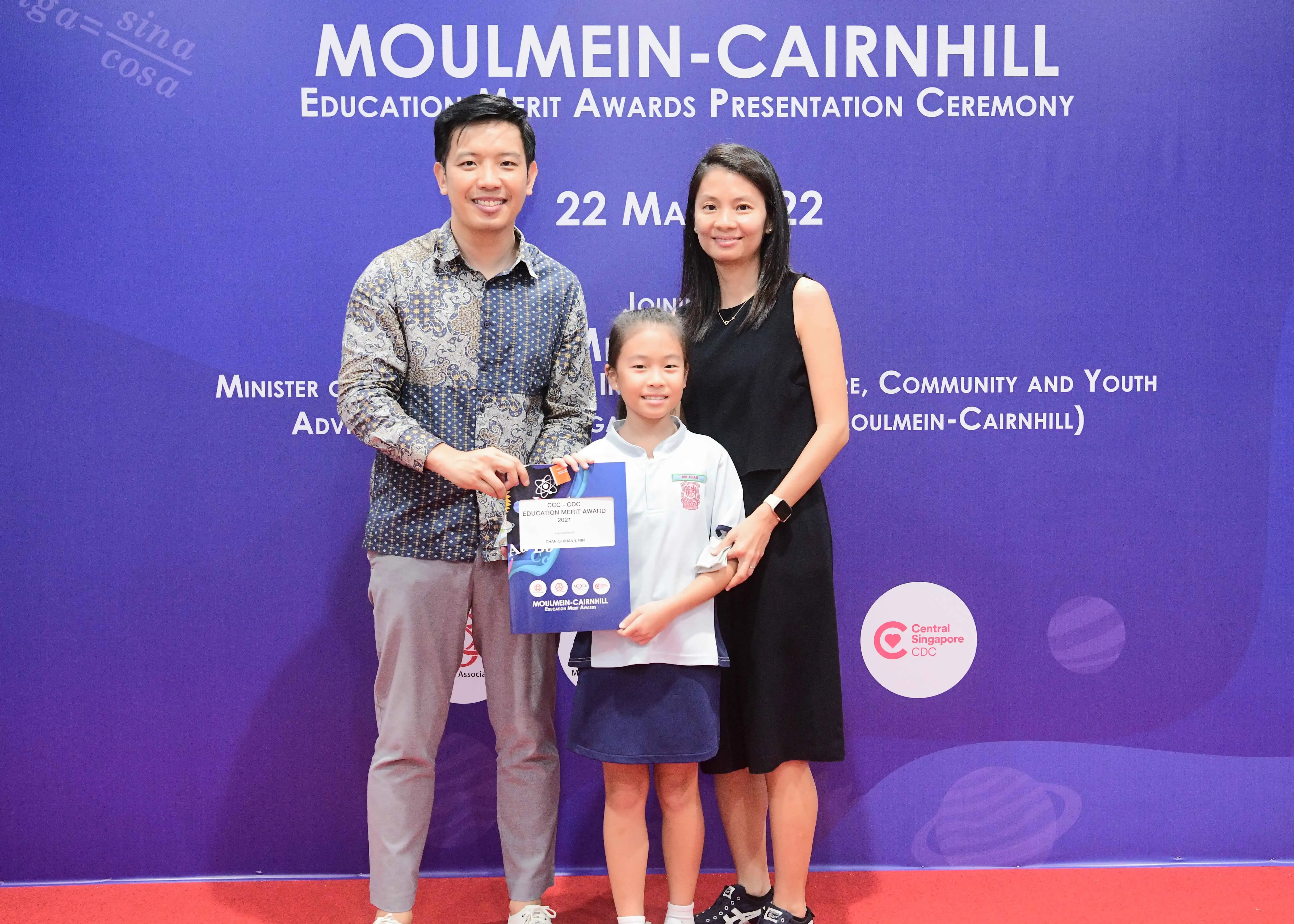 Two adults and a young girl pose with an award certificate at the Moulmein-Cairnhill Education Merit Awards ceremony.