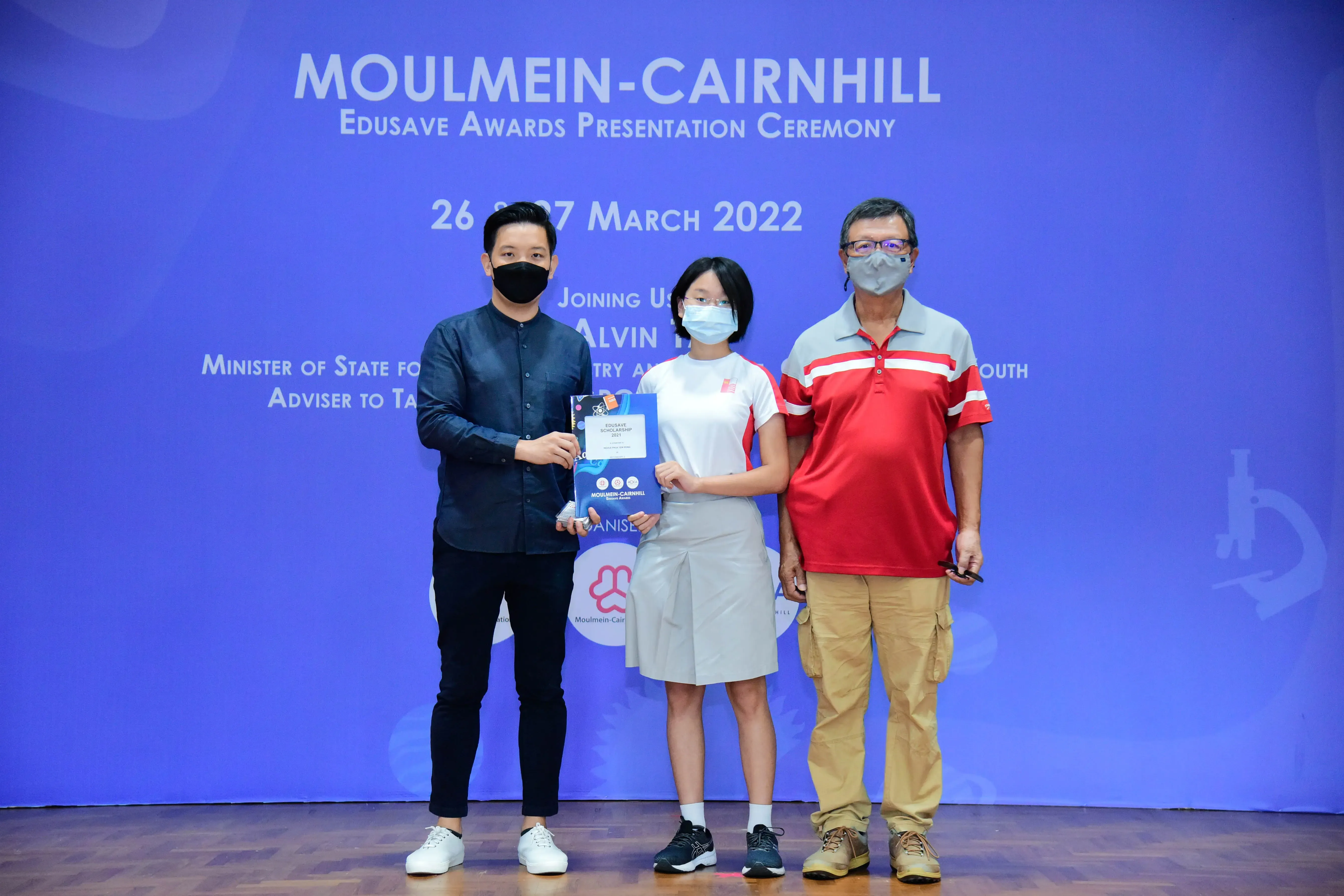 Three masked people stand before a blue Moulmein-Cairnhill Edusave Awards Presentation Ceremony backdrop, center holds an award.