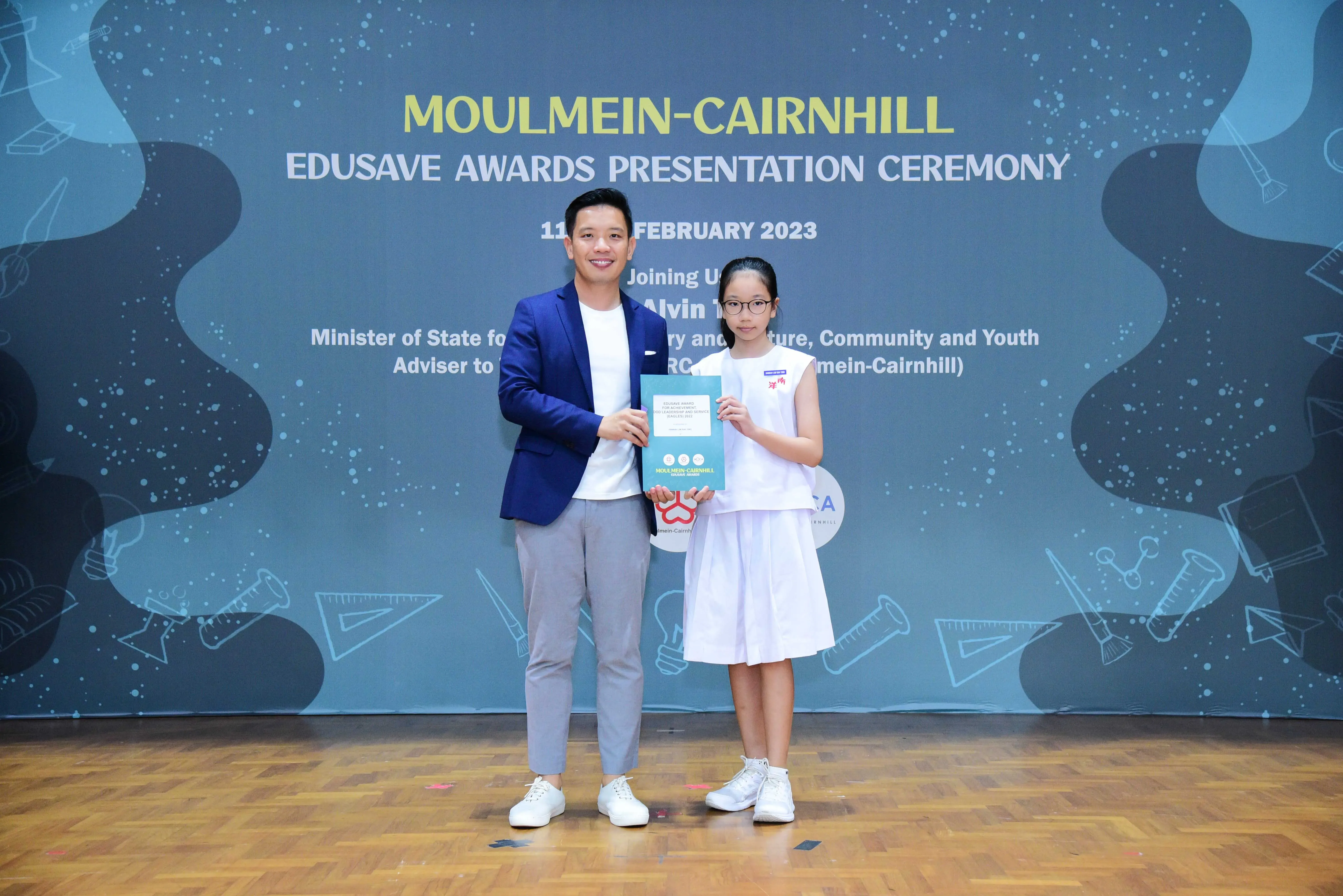 A man in a blue blazer and a young girl in white school uniform hold a certificate at the Moulmein-Cairnhill Edusave Awards 2023.