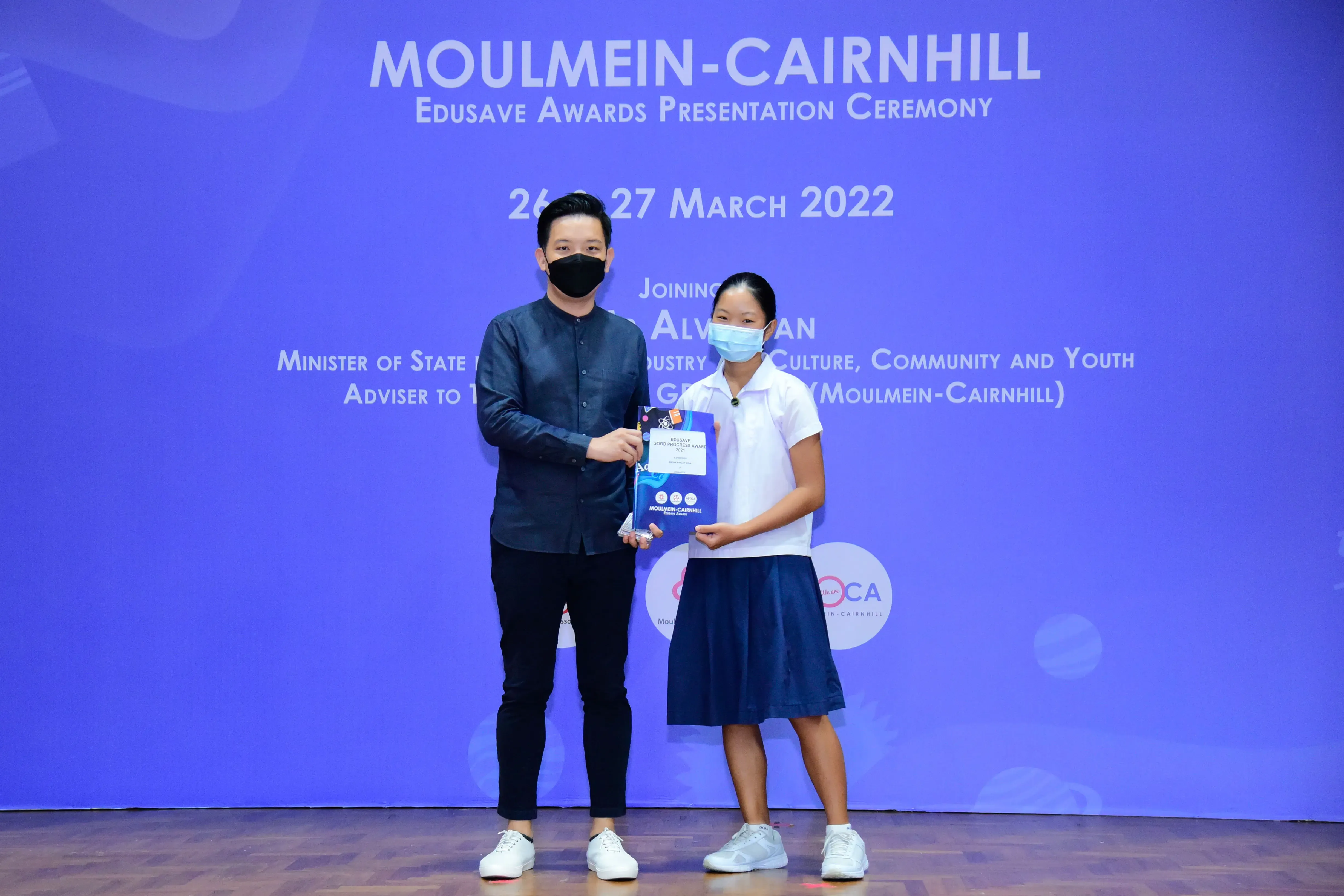 A masked man and a young girl in school uniform hold an award at the Moulmein-Cairnhill Edusave Awards Ceremony 2022.
