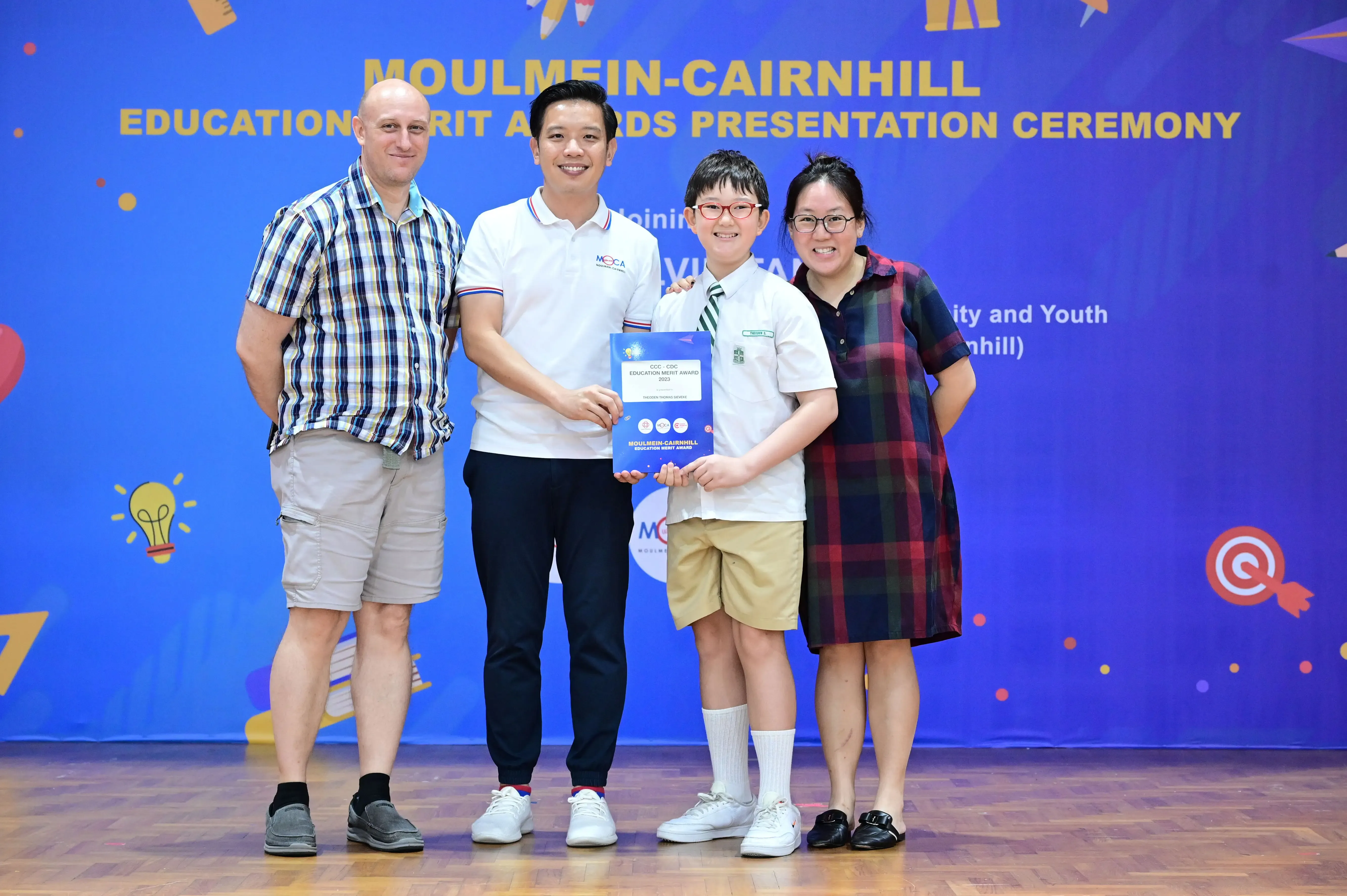 Four people pose at the Moulmein-Cairnhill Education Merit Awards Presentation Ceremony, with a child holding a certificate.