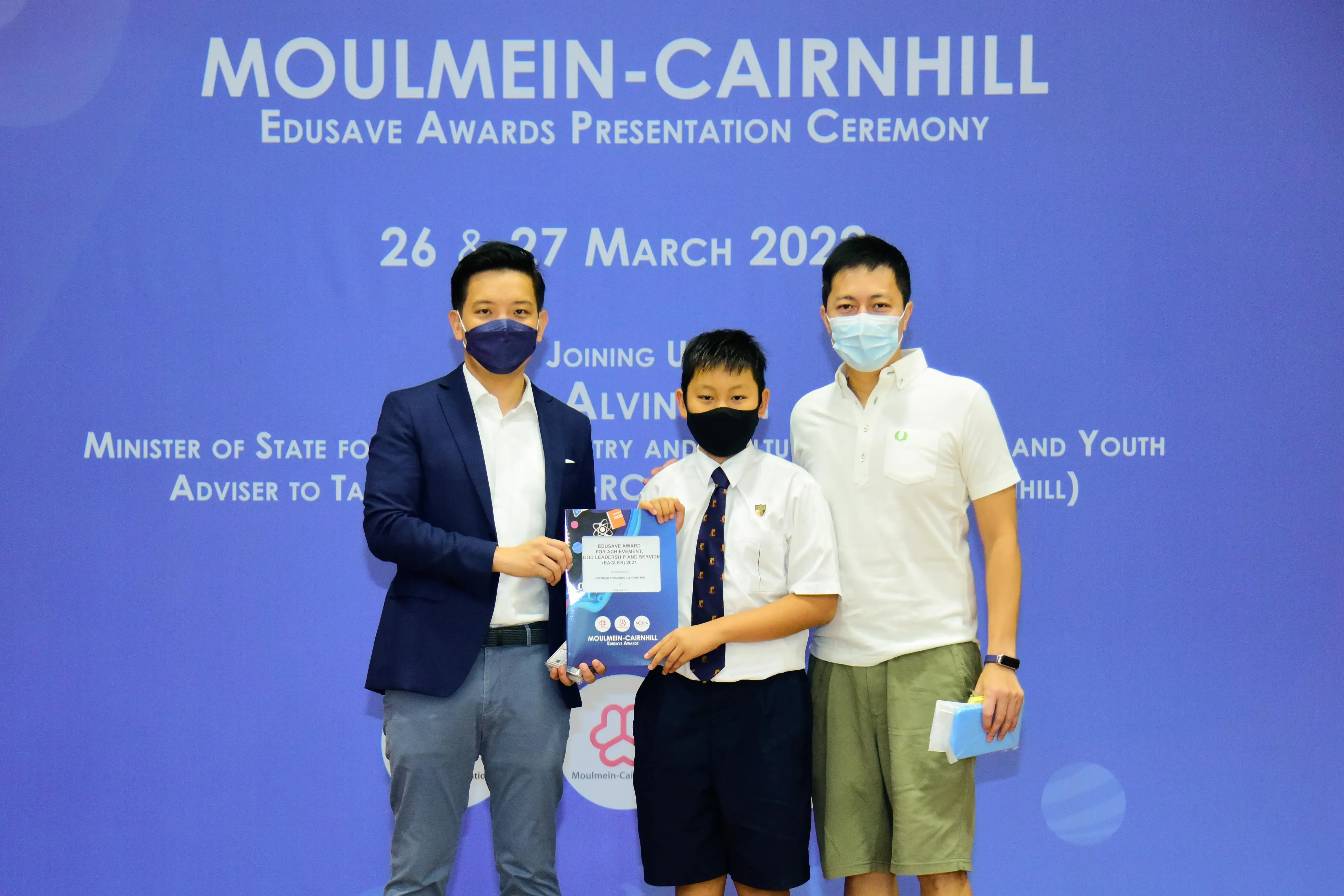 Three masked people pose at the Moulmein-Cairnhill Edusave Awards Presentation Ceremony, with a child holding an award.