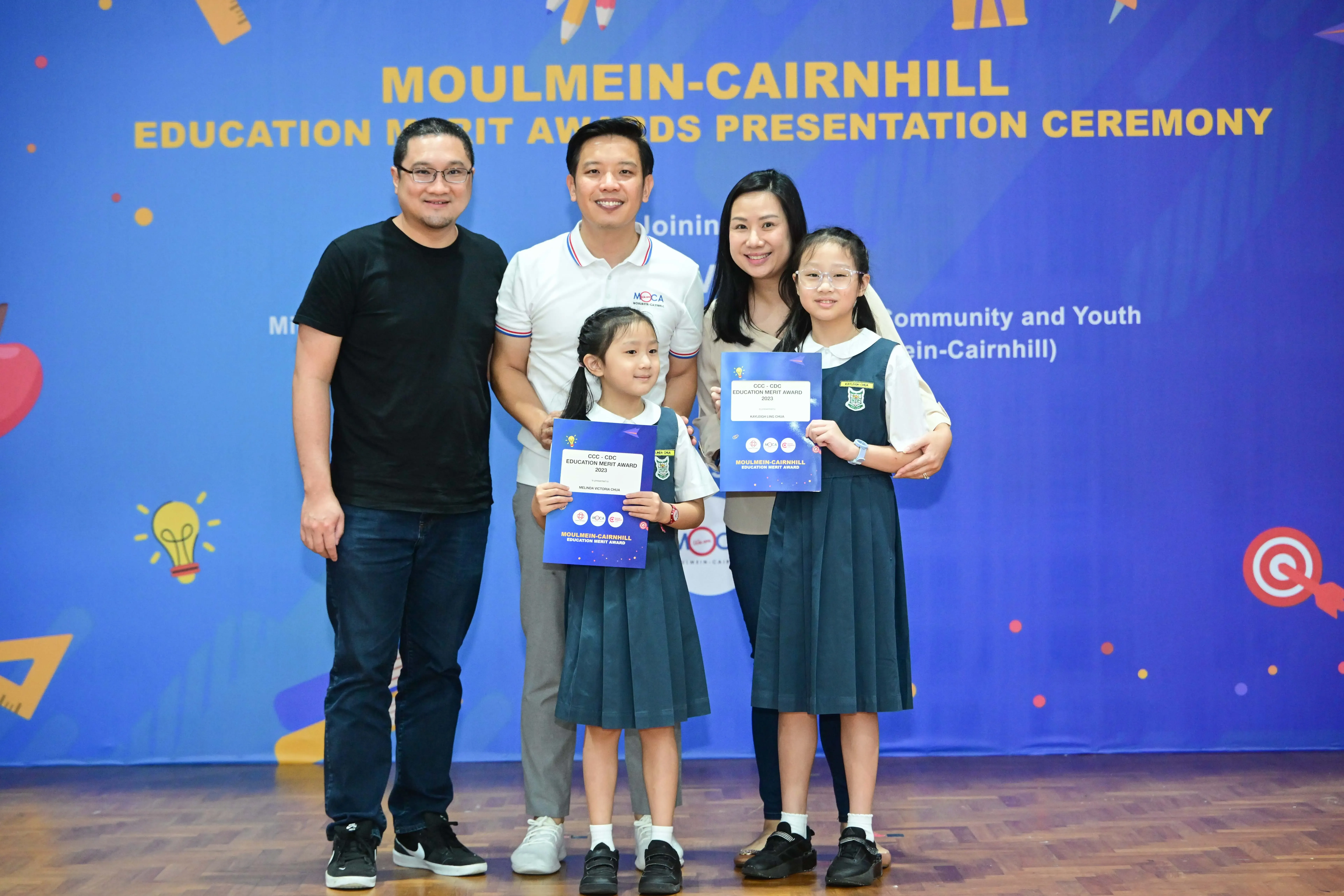 Two adults and two girls in school uniforms hold award certificates at the Moulmein-Cairnhill Education Merit Awards ceremony.