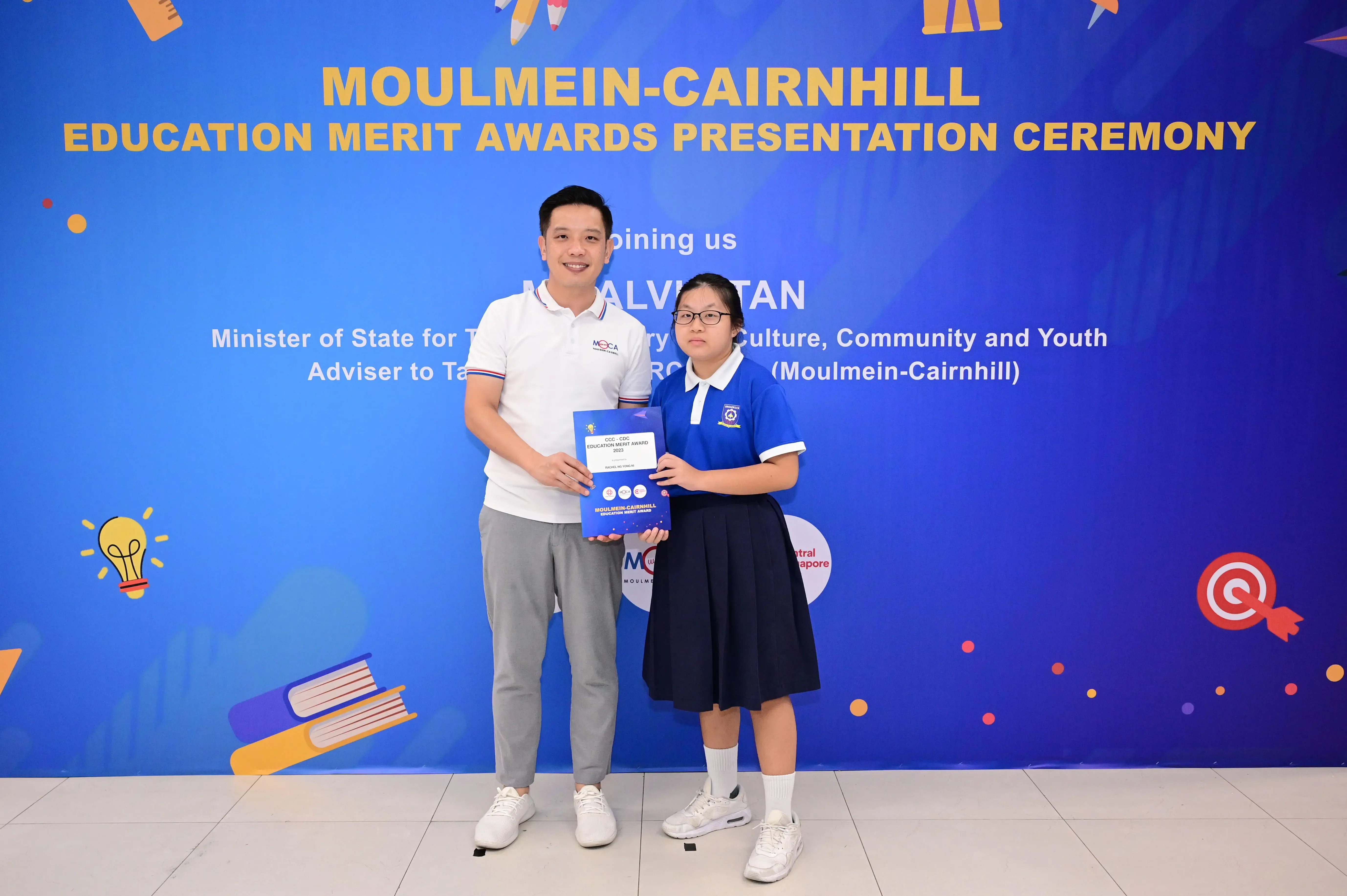 A man and a young student hold an award certificate in front of a Moulmein-Cairnhill Education Merit Awards banner.