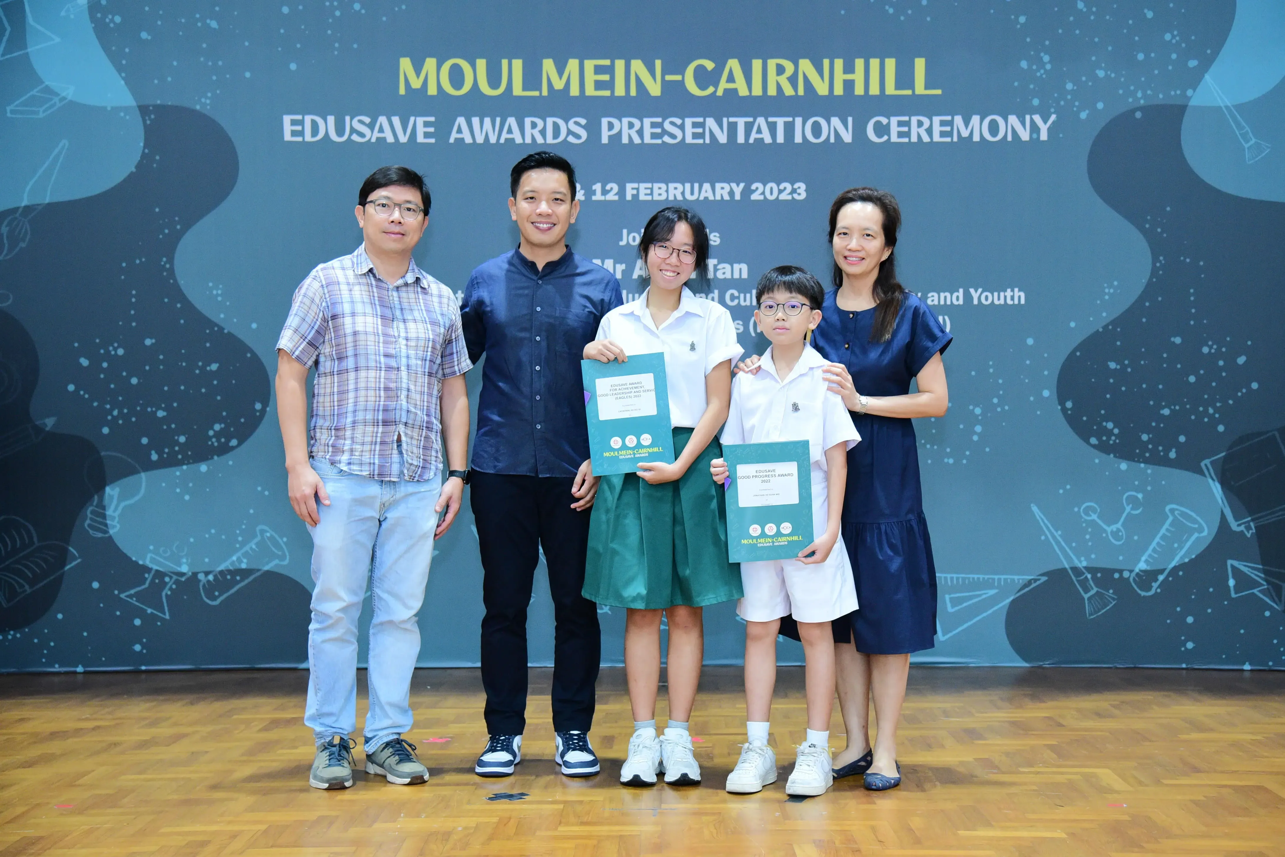Five people pose at the Moulmein-Cairnhill Edusave Awards Presentation Ceremony, with two children holding award folders.