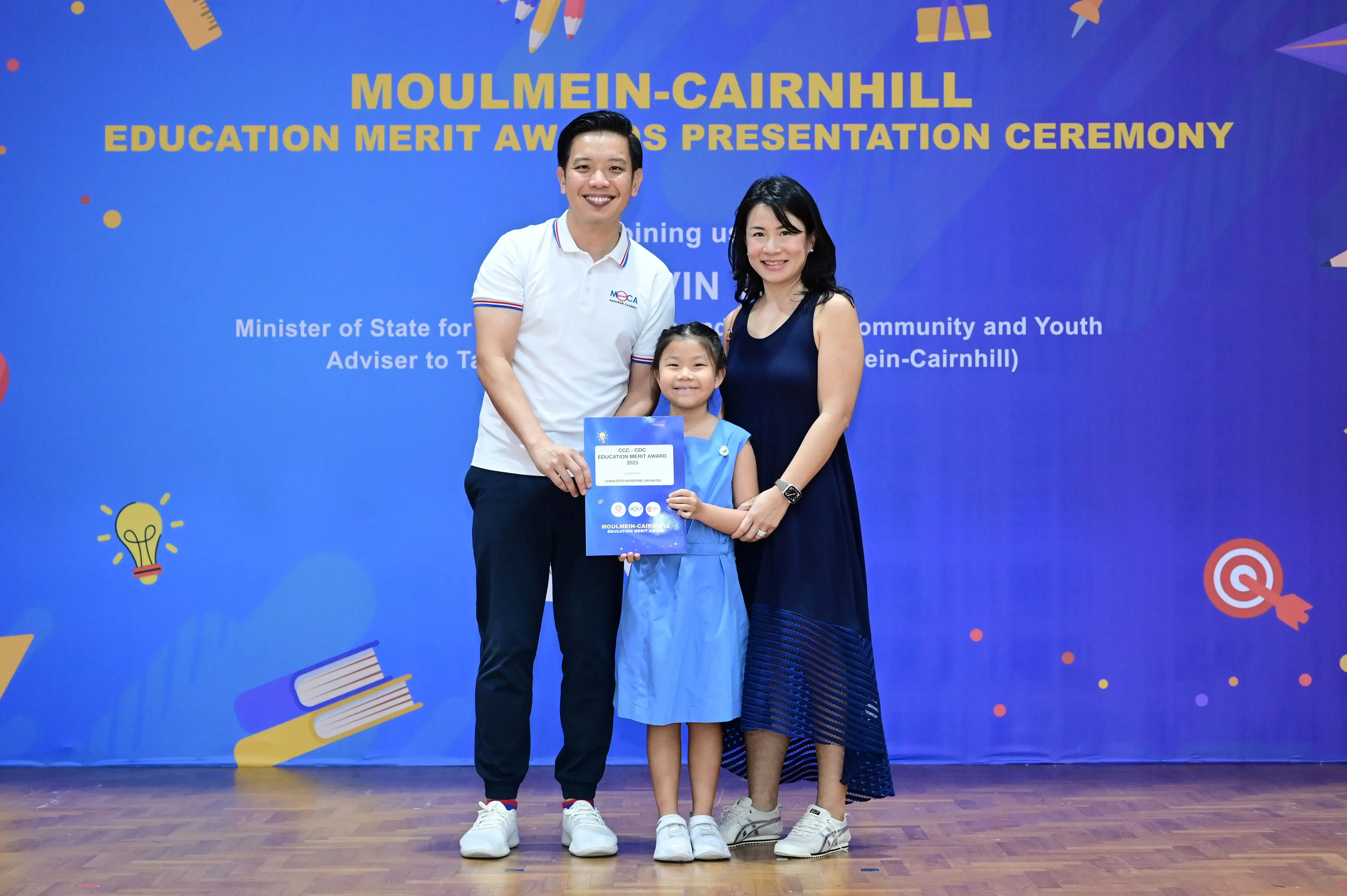 A young girl holds an award certificate flanked by two adults at the Moulmein-Cairnhill Education Merit Awards Ceremony.